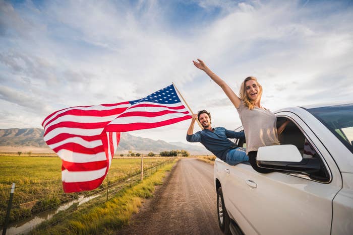 Two people joyfully lean out of a car with an American flag, set against a scenic rural landscape on a clear day