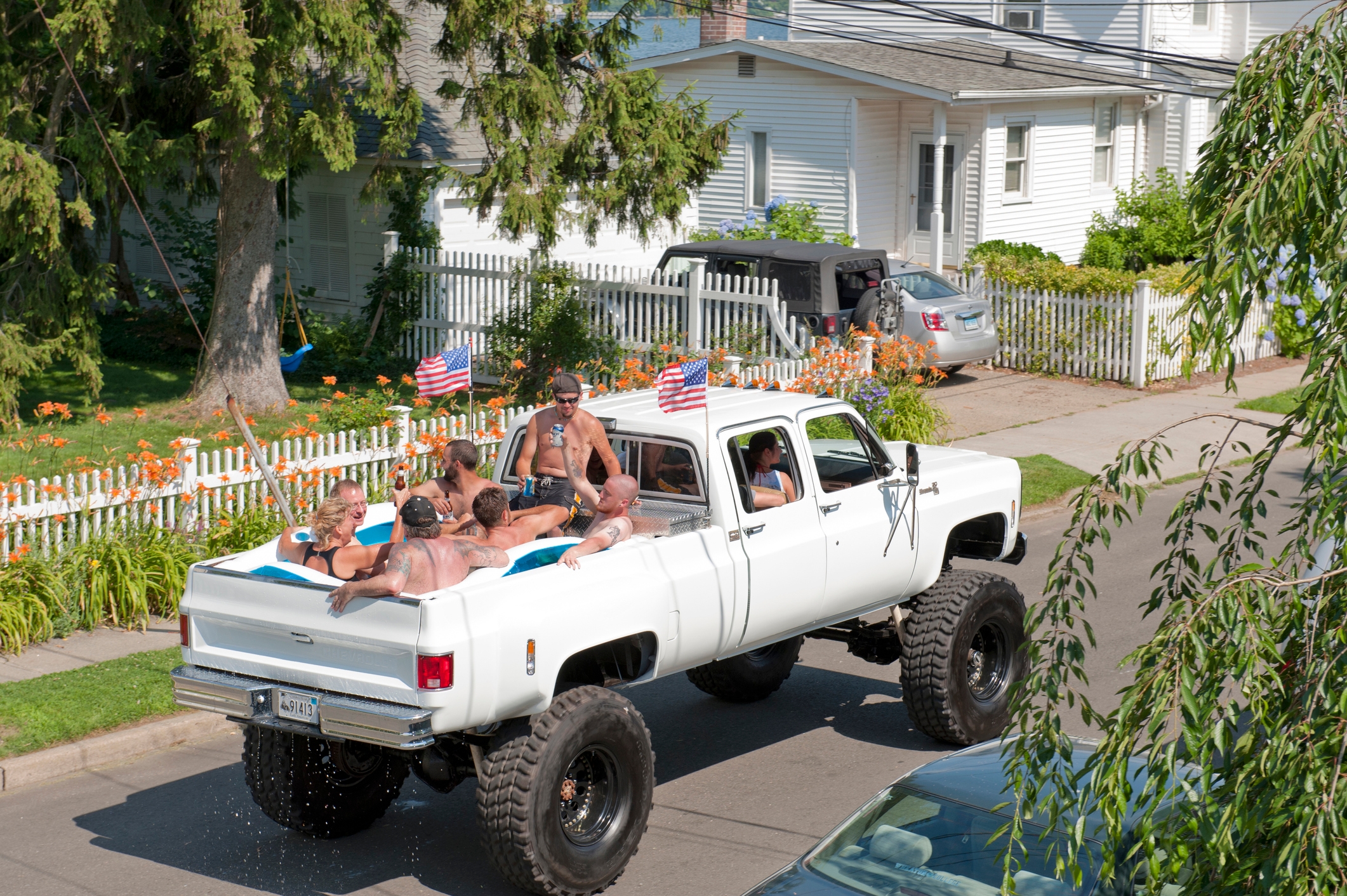 A group of people are sitting in a pool in the bed of a raised pickup truck driving down a residential street. Two small American flags are visible