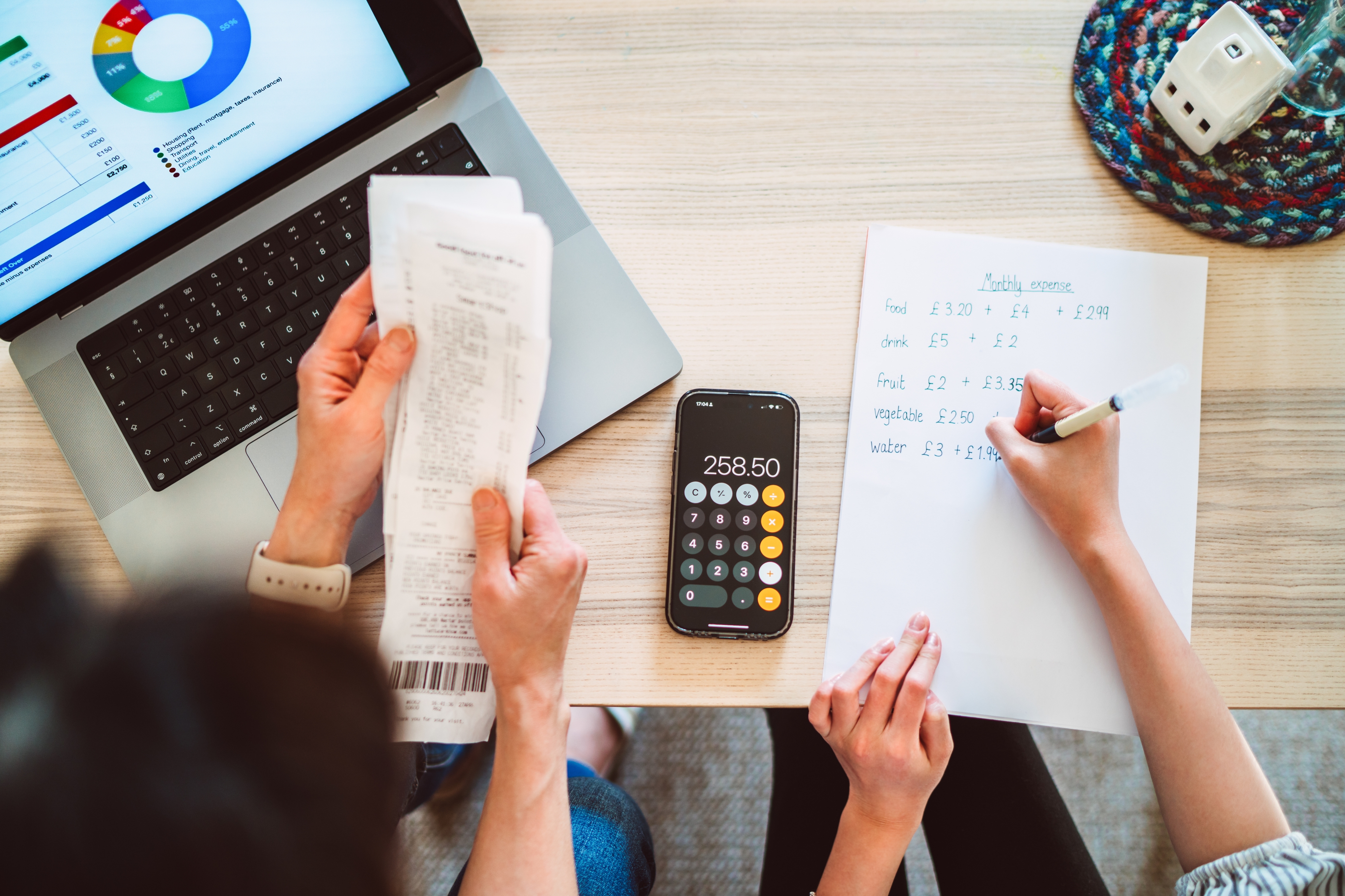 Person holding a receipt while another writes expenses on paper, with a phone calculator showing 258.50 on a table next to a laptop