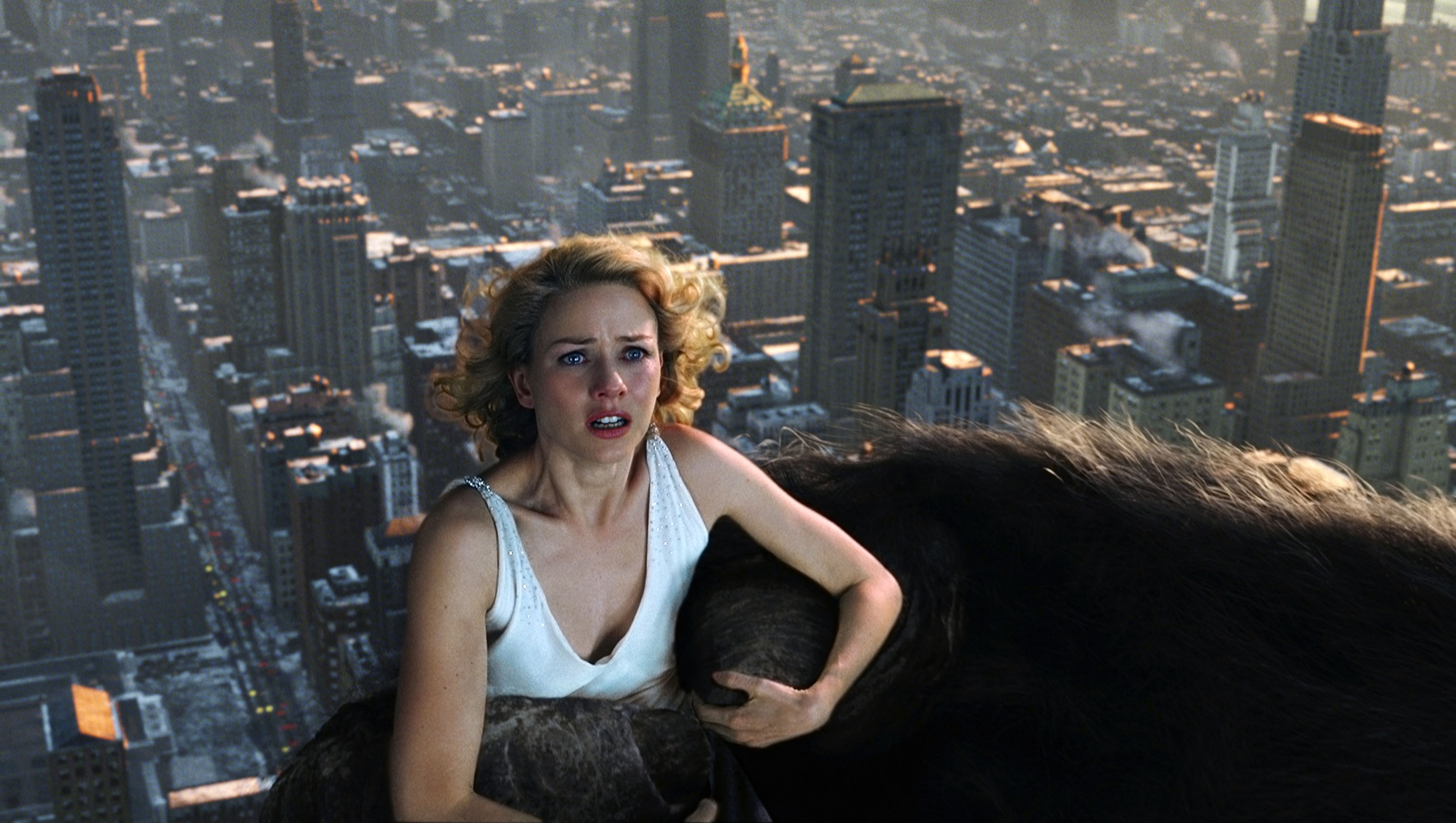 Person in a white tank top clings to King Kong's arm atop a cityscape, expressing fear and awe