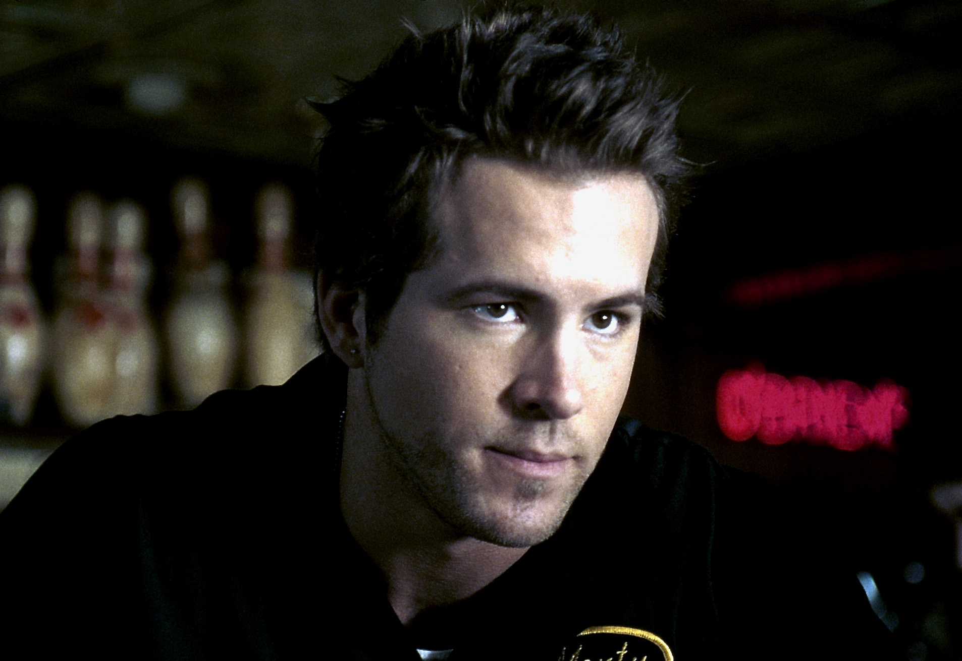 Man in a casual black shirt with bowling pins in the background, appearing focused, in a scene potentially from a film or TV show