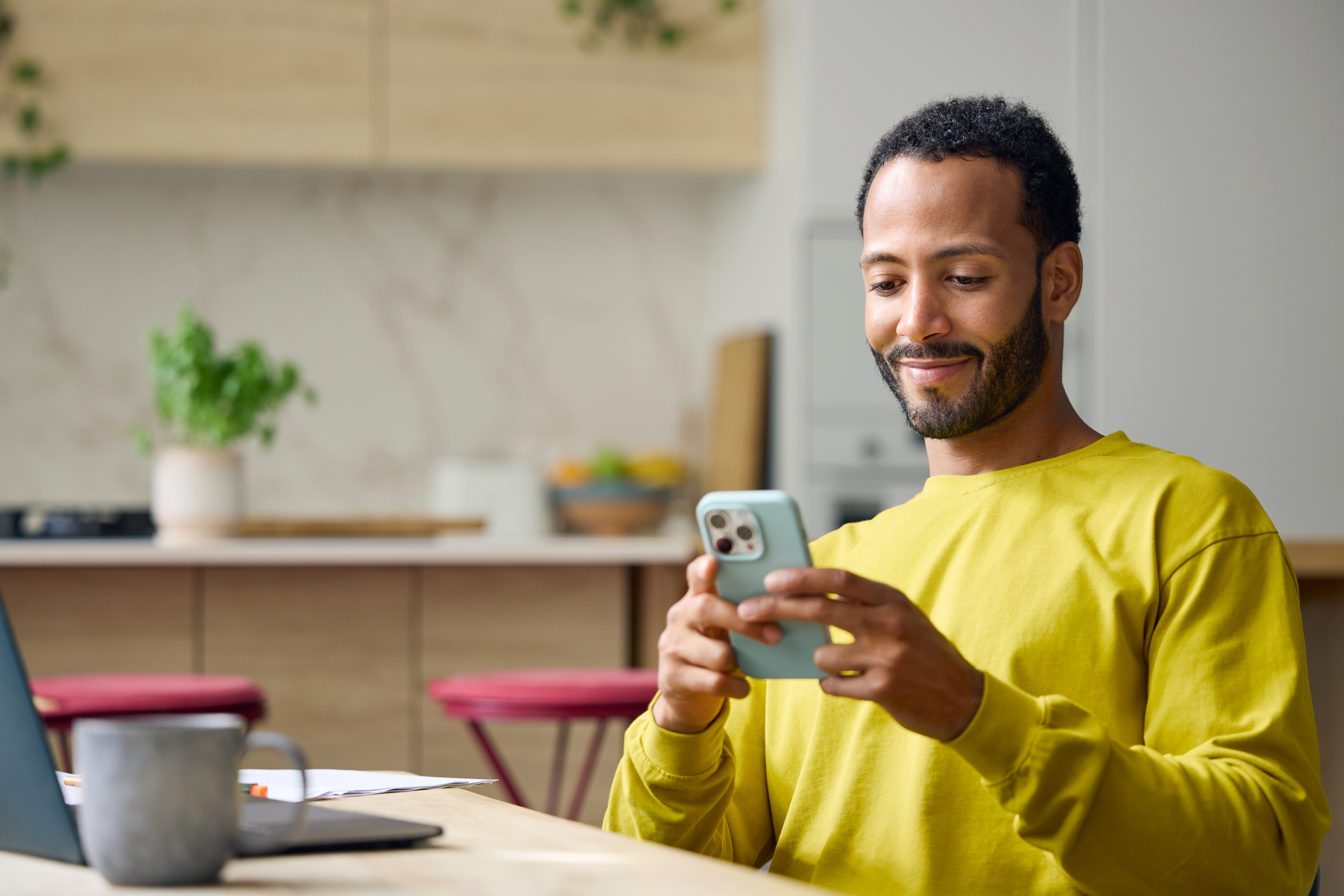 Man in a cozy kitchen smiling at his phone, sitting at a table with a laptop and a cup