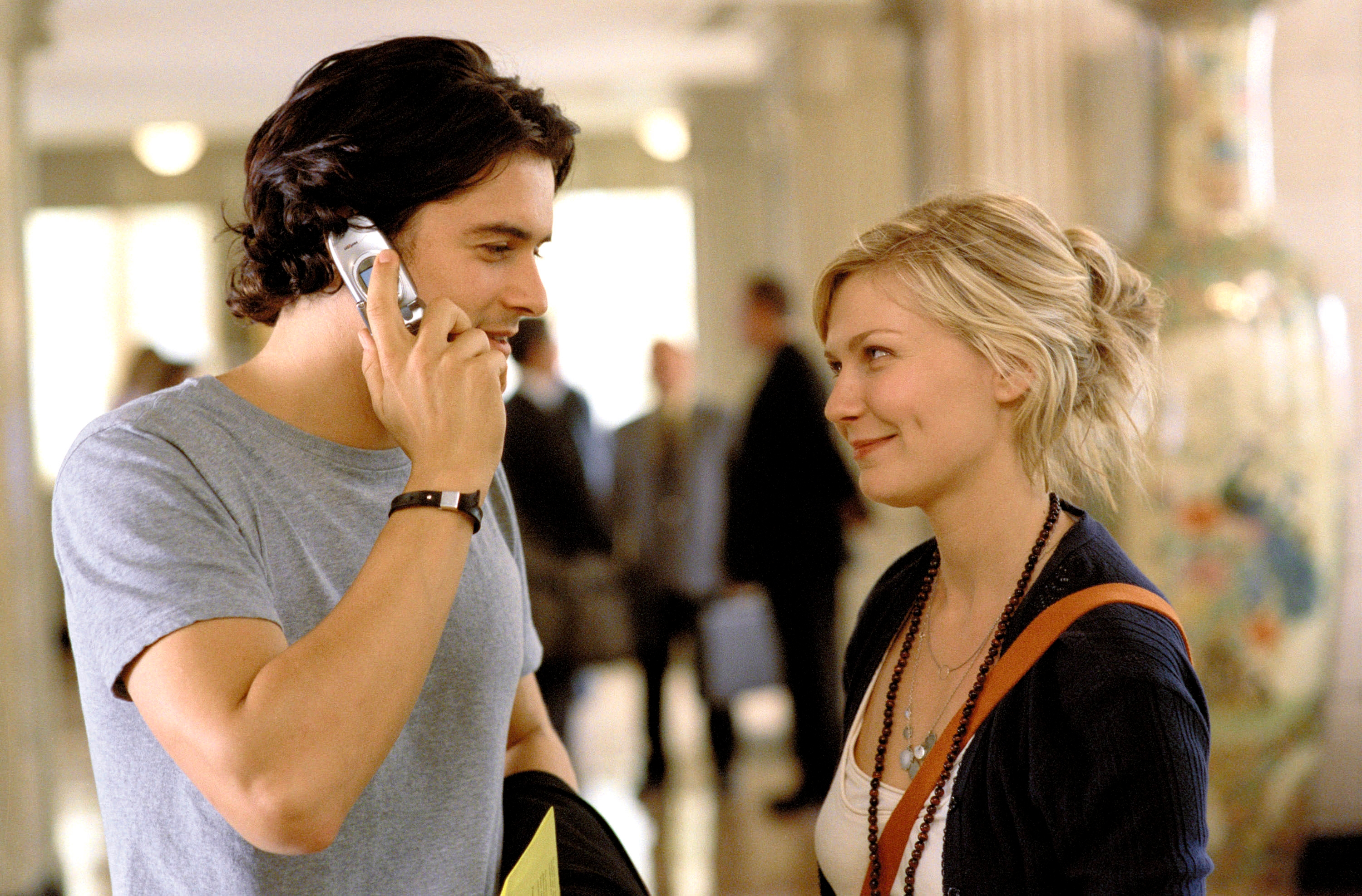 A man on the phone holds a letter, standing next to a smiling woman. Both are casually dressed, interacting in a public indoor setting