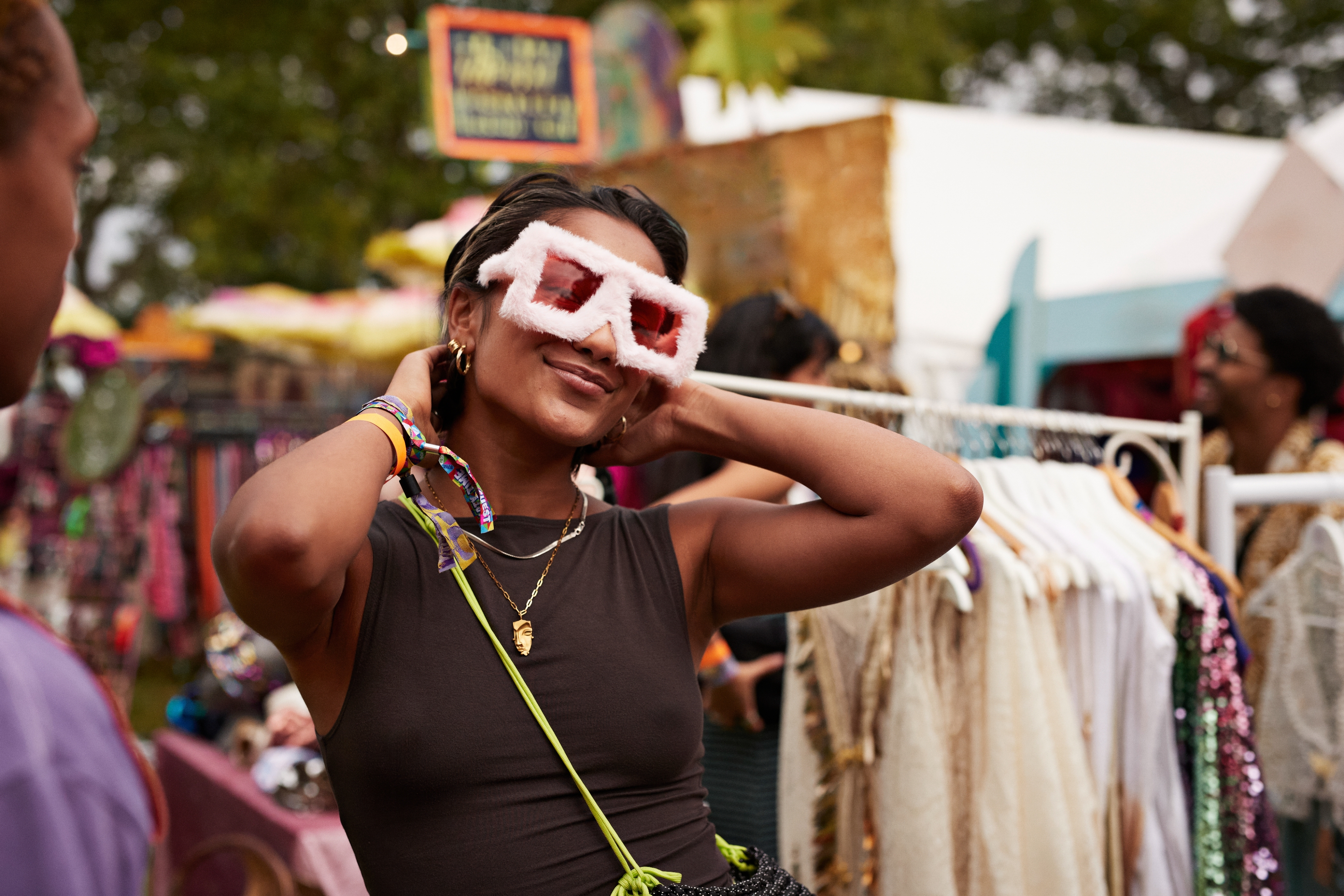 Person smiling and adjusting oversized fuzzy glasses at an outdoor market, surrounded by colorful booths and clothing racks