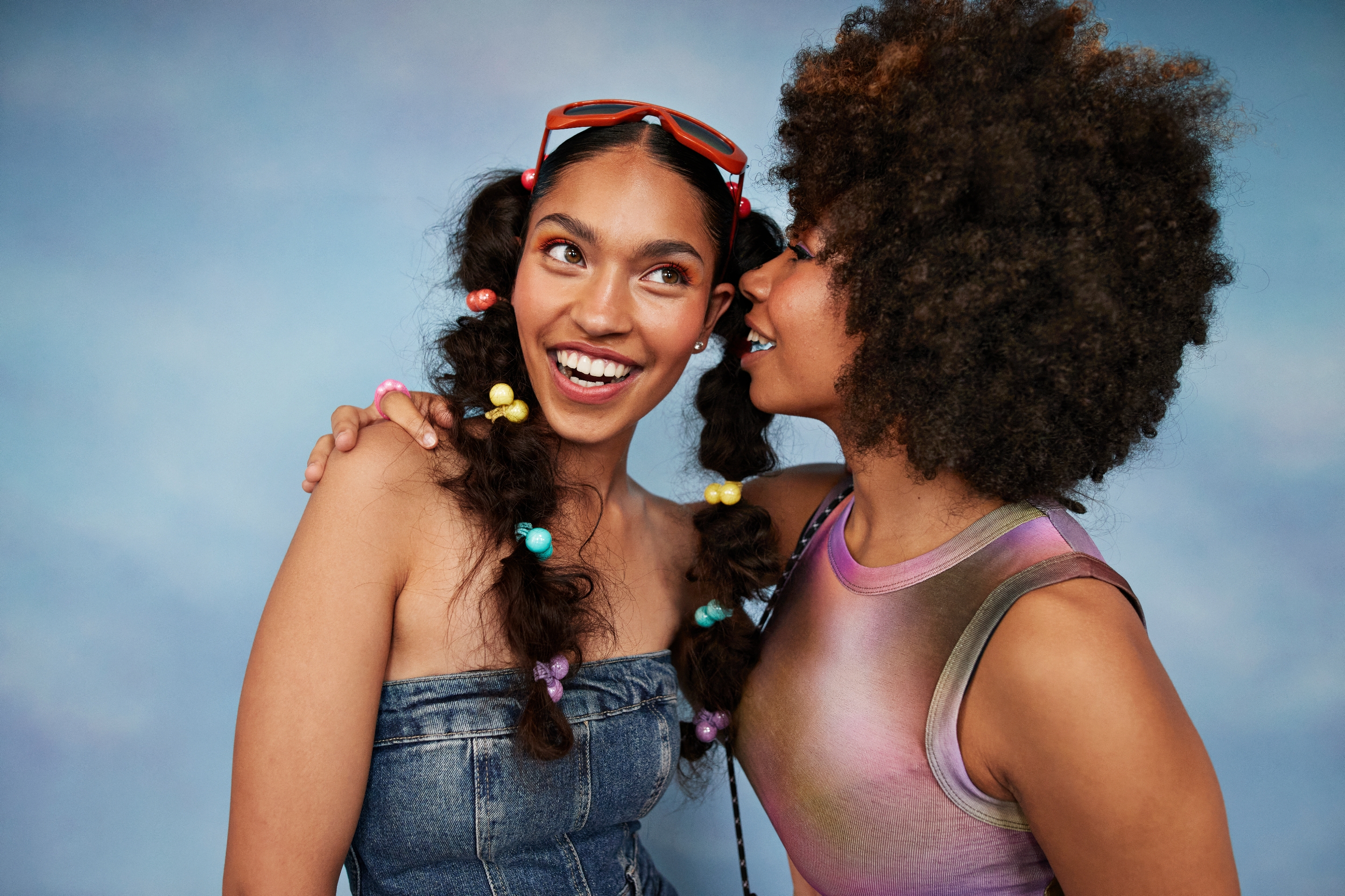 Two people smiling; one whispers to the other. The left person wears a denim outfit with colorful hair ties, the right wears a sleeveless top