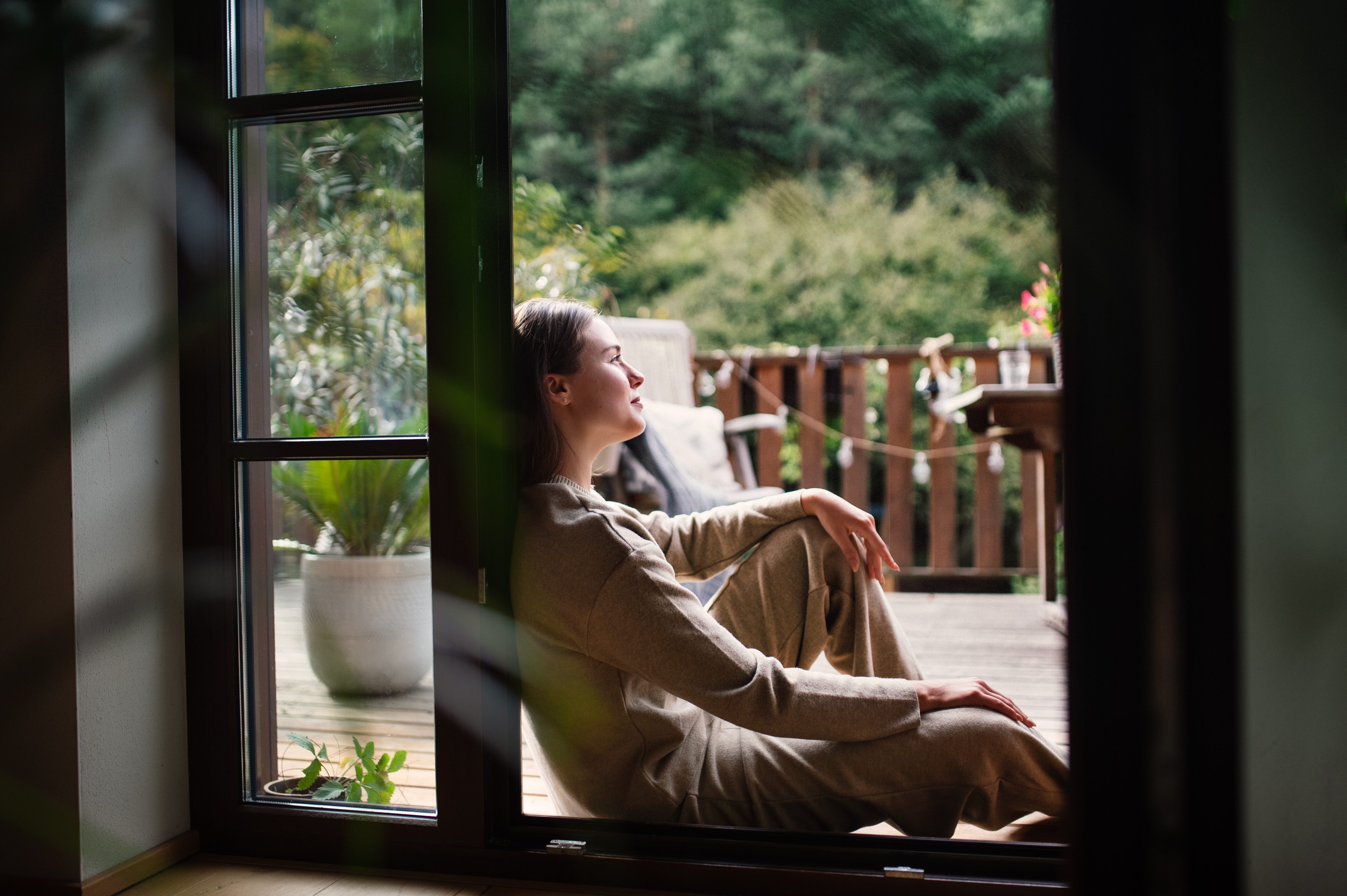 Person sitting in a doorway, gazing outside, dressed in relaxed, casual attire. A peaceful outdoor setting visible beyond the deck