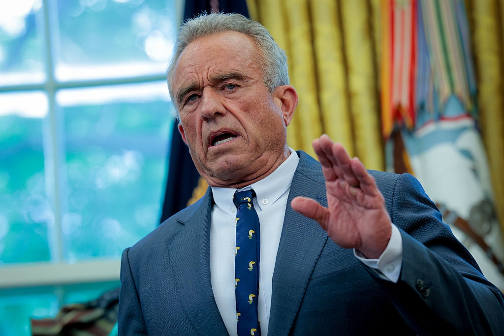 Person in a suit and tie stands indoors, gesturing with one hand, appearing to speak or address an audience