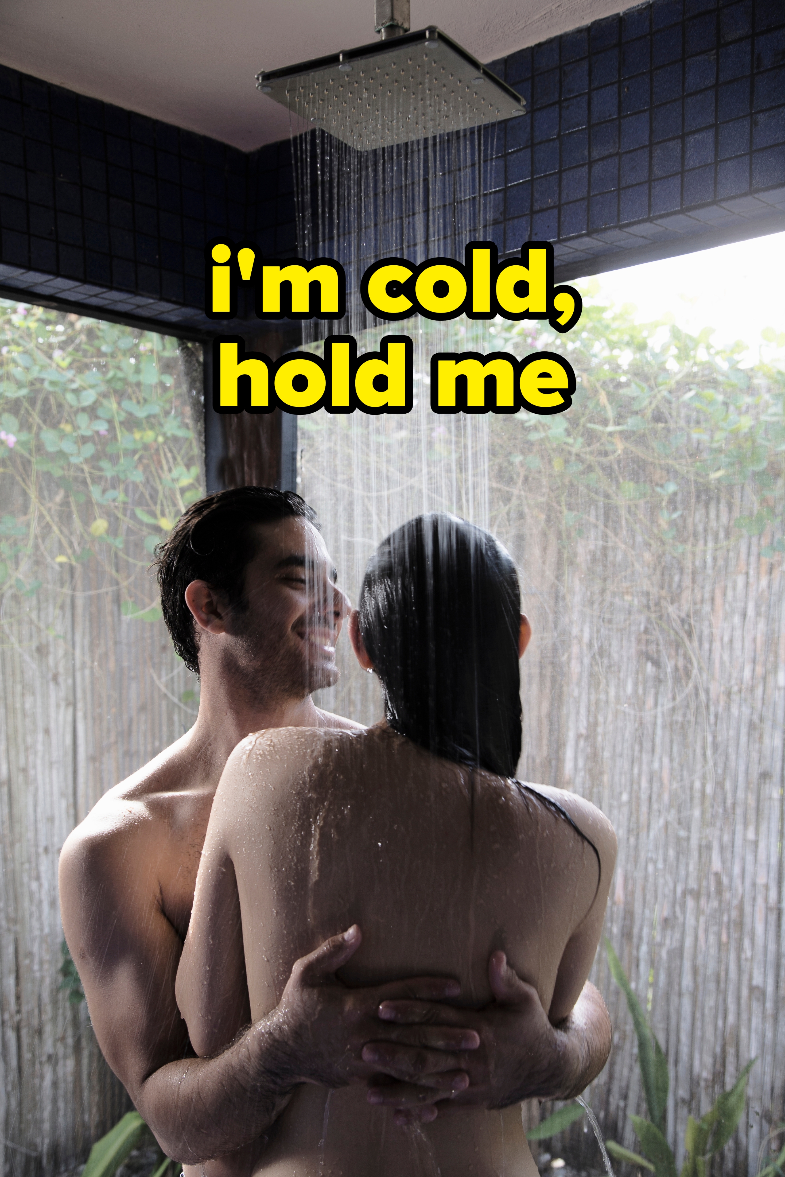 Two people embracing under an outdoor shower, smiling at each other. The setting is private, with greenery visible in the background
