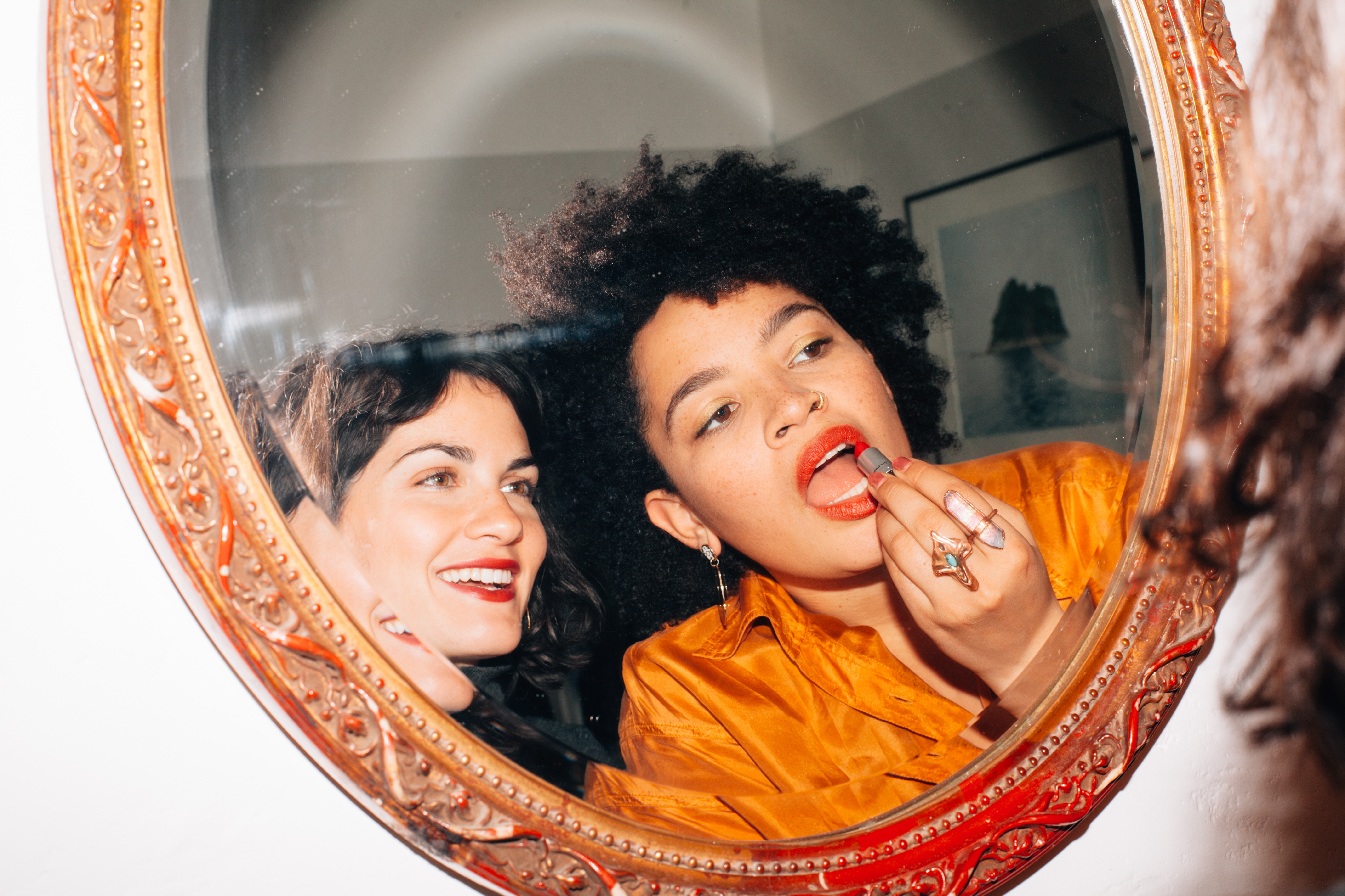 Two people smile as they apply lipstick in a mirror. One wears a shiny blouse, while the other sports a casual look