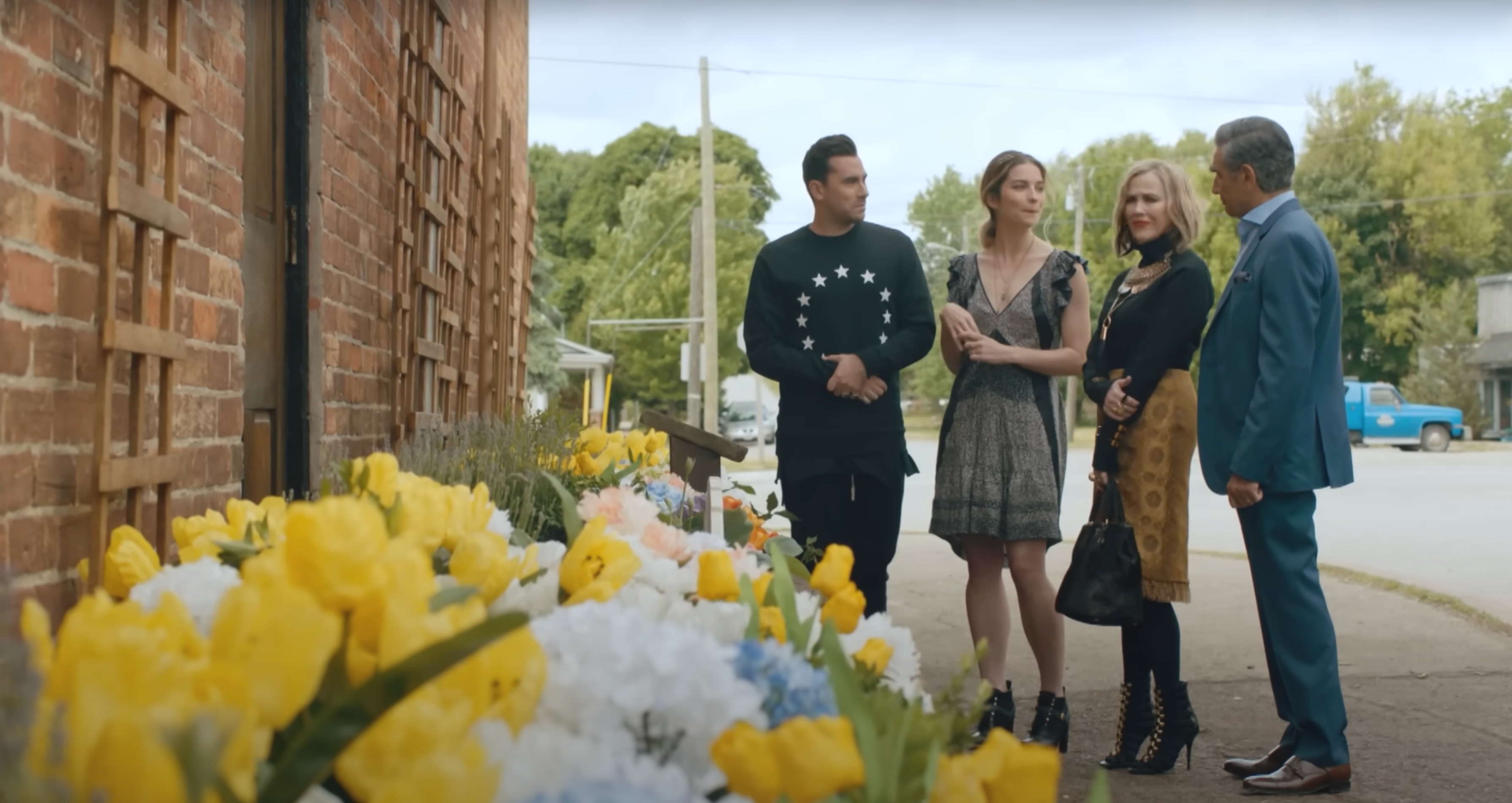 Four people stand on a street near a brick wall with flowers. They appear engaged in conversation, dressed in casual and stylish outfits