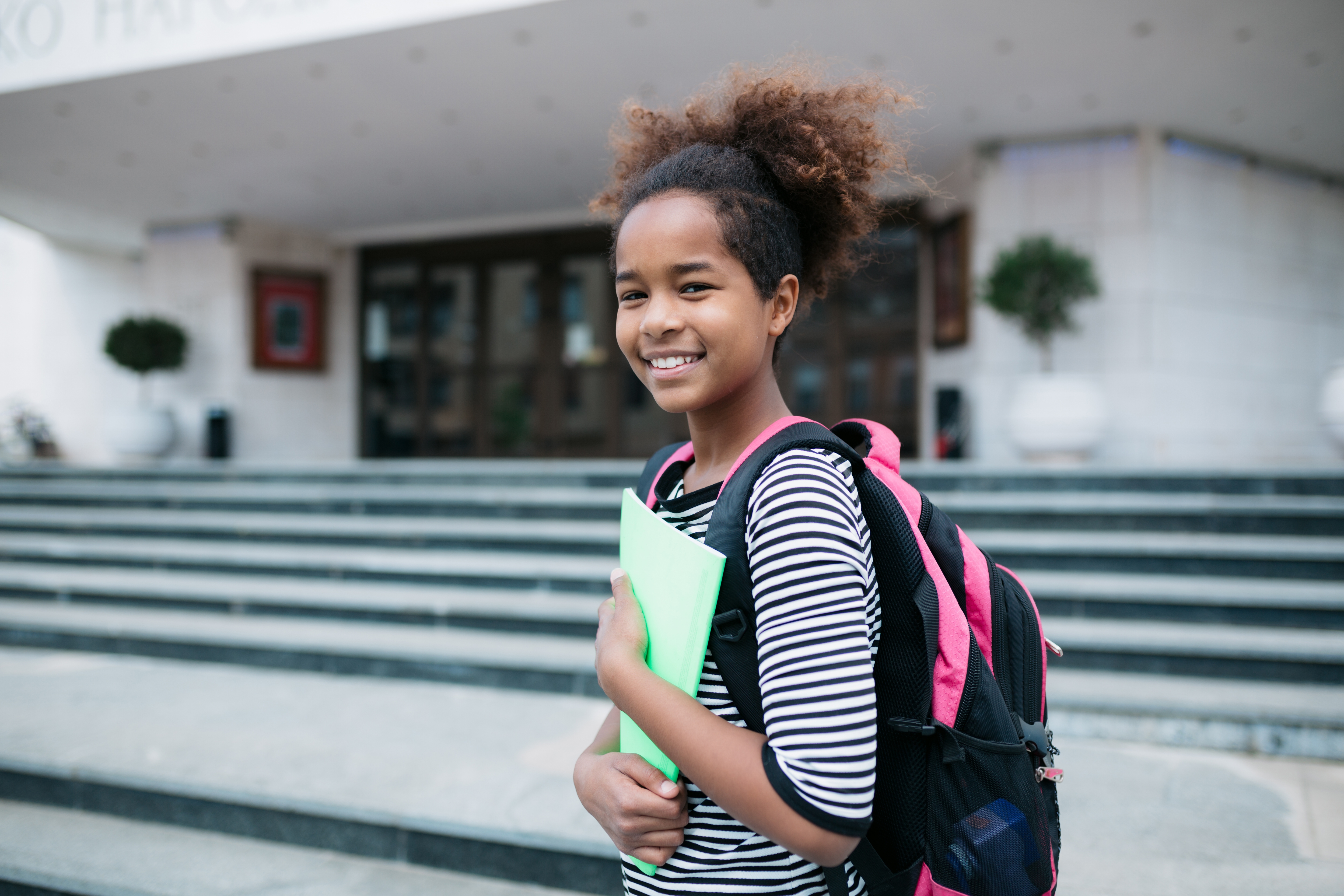 Smiling student with a backpack holds a folder, standing in front of school steps