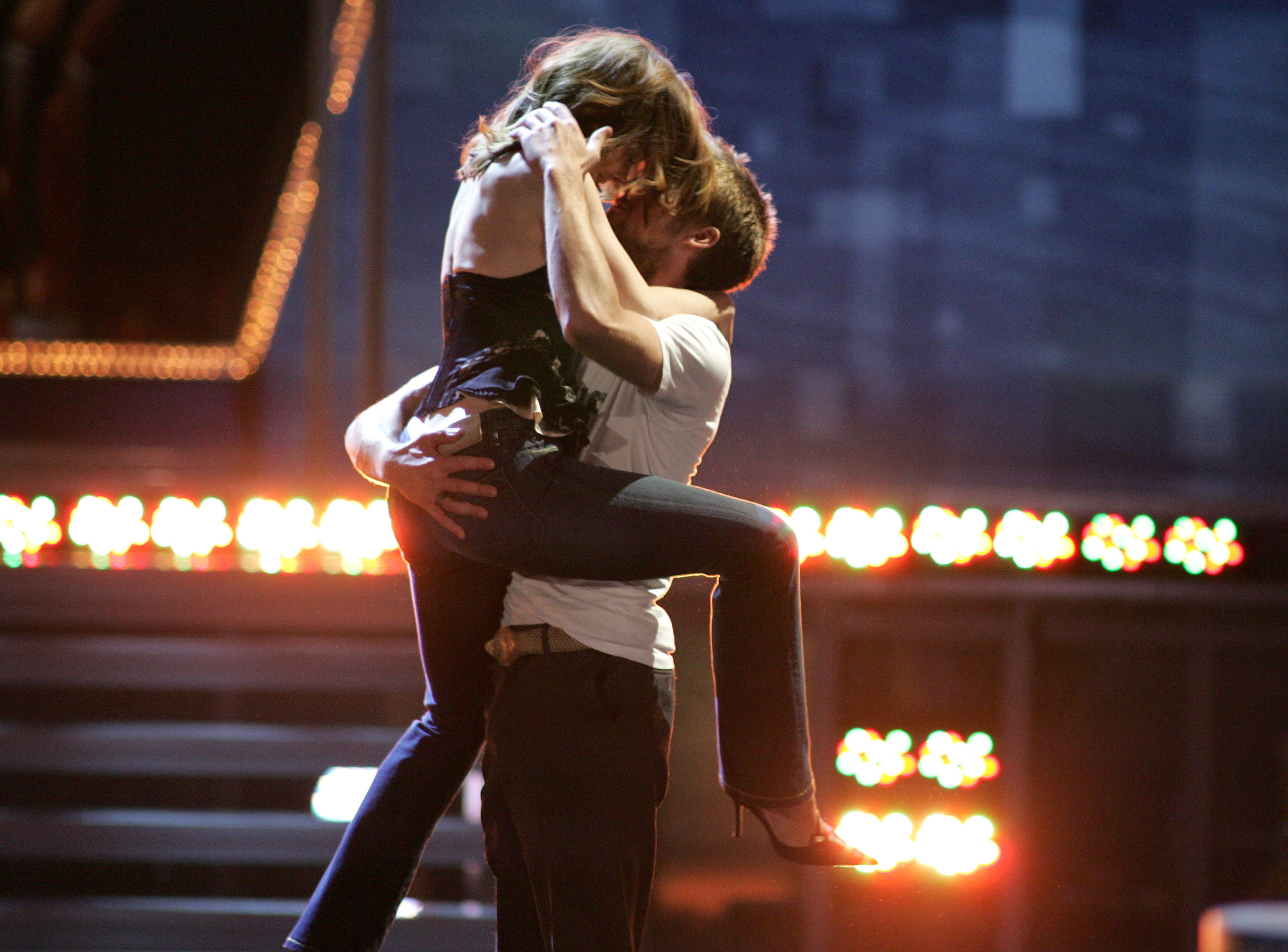 A couple passionately embraces on stage during a performance. One person is lifted into the air, both are in casual attire