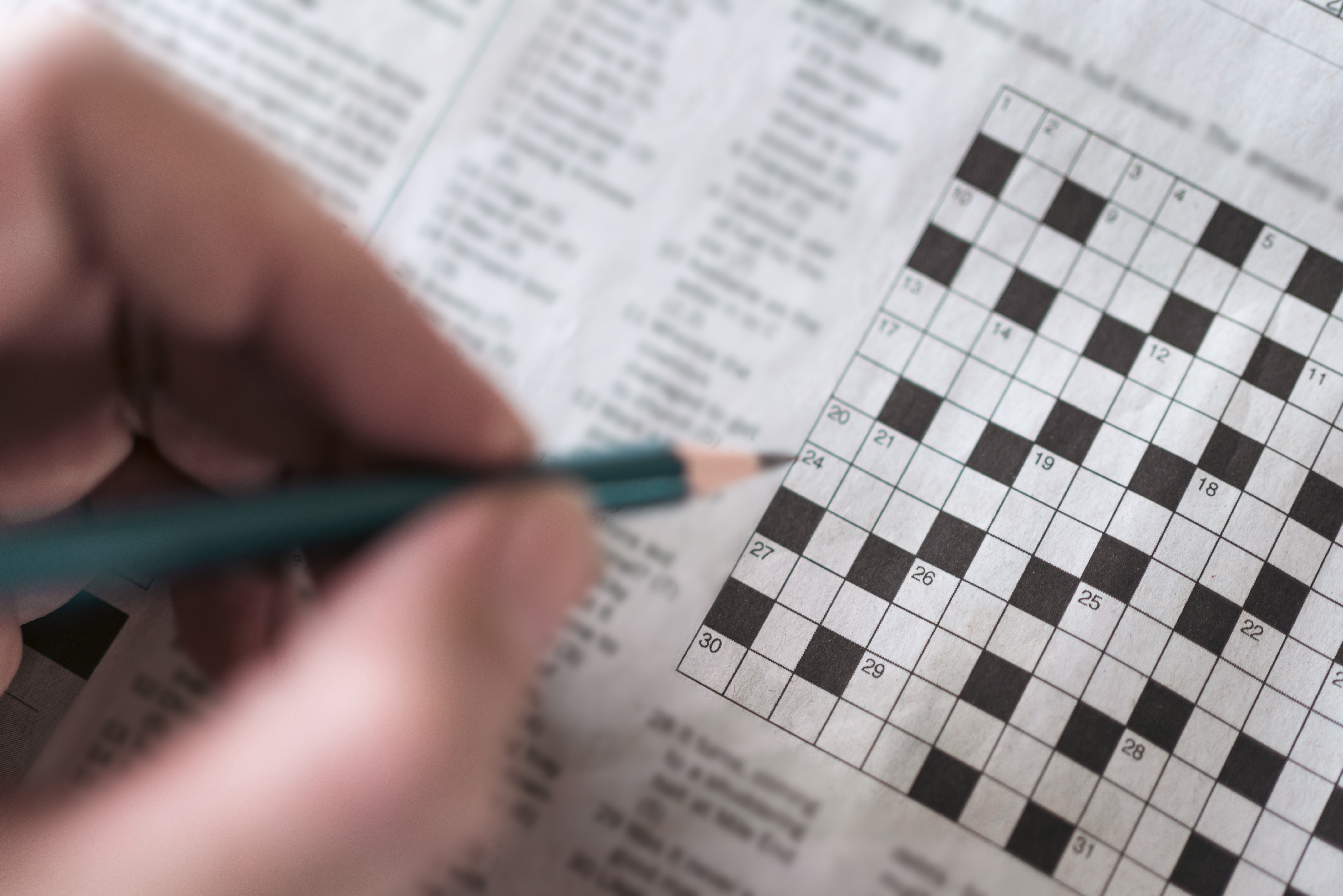 A person holds a pencil over a partially completed newspaper crossword puzzle, suggesting a moment of leisure and mental engagement