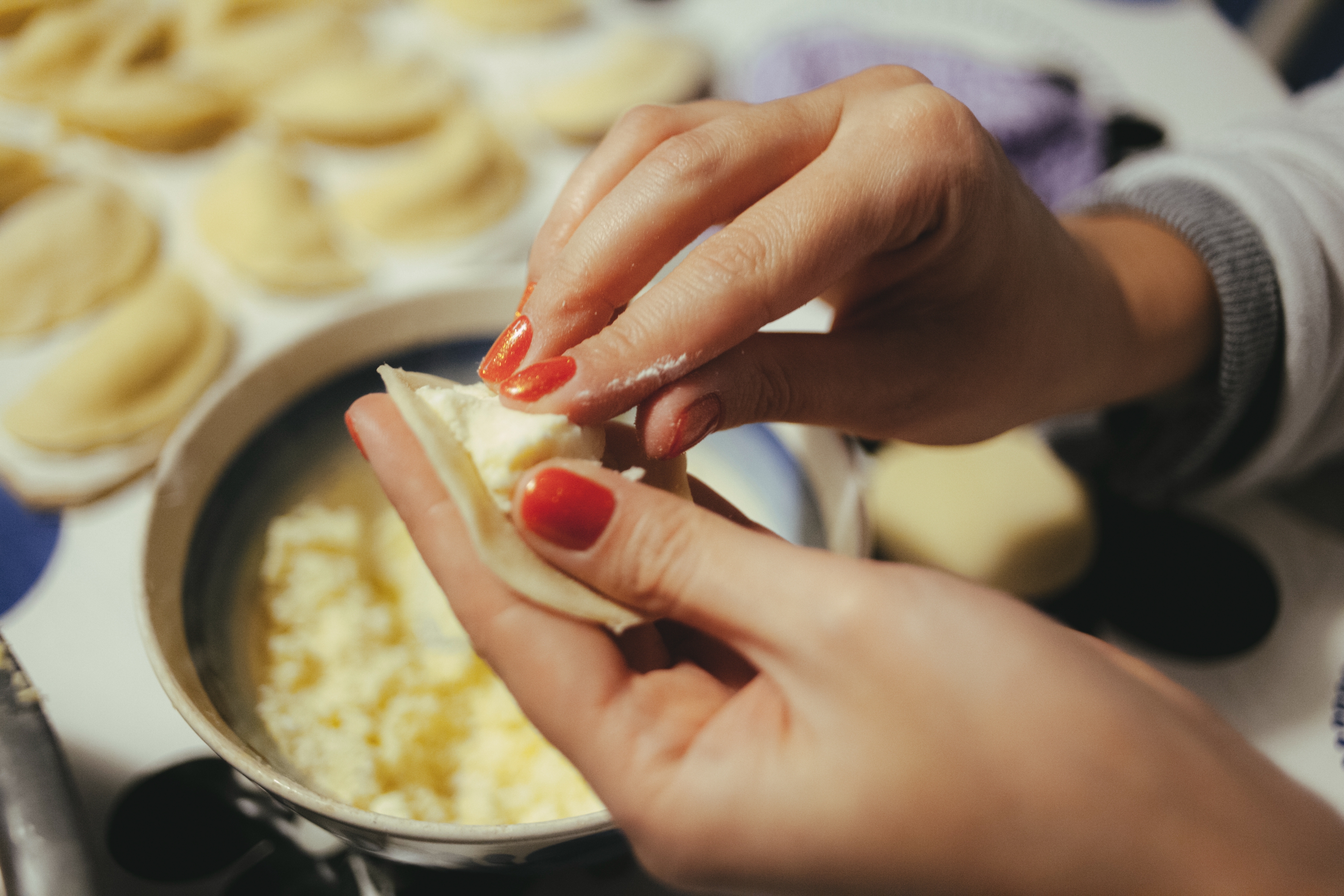 Hands fill dumplings with cheese mixture in a kitchen setting, with more dumplings prepared on the counter