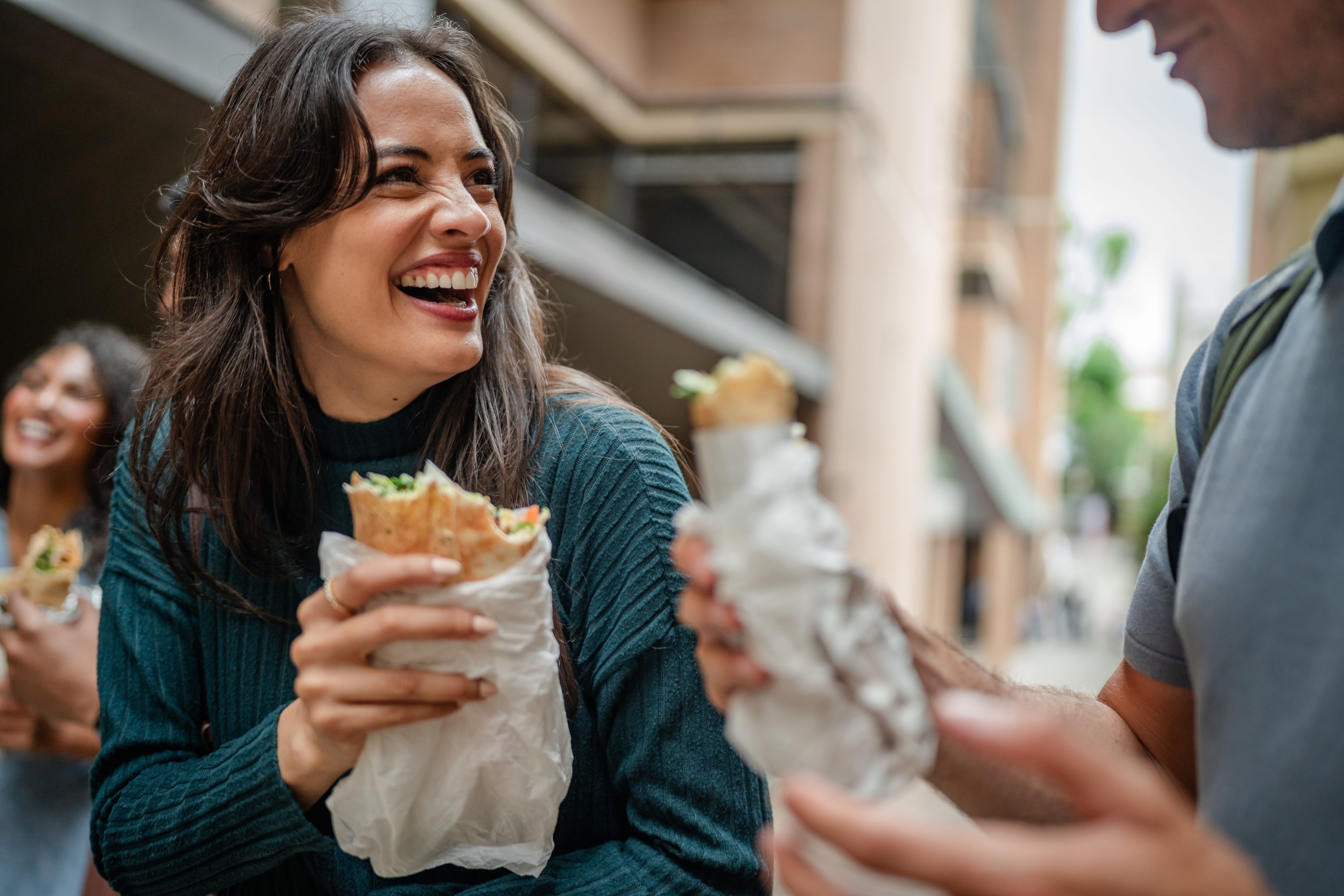 Three people enjoy wrapped sandwiches outdoors, smiling and conversing casually