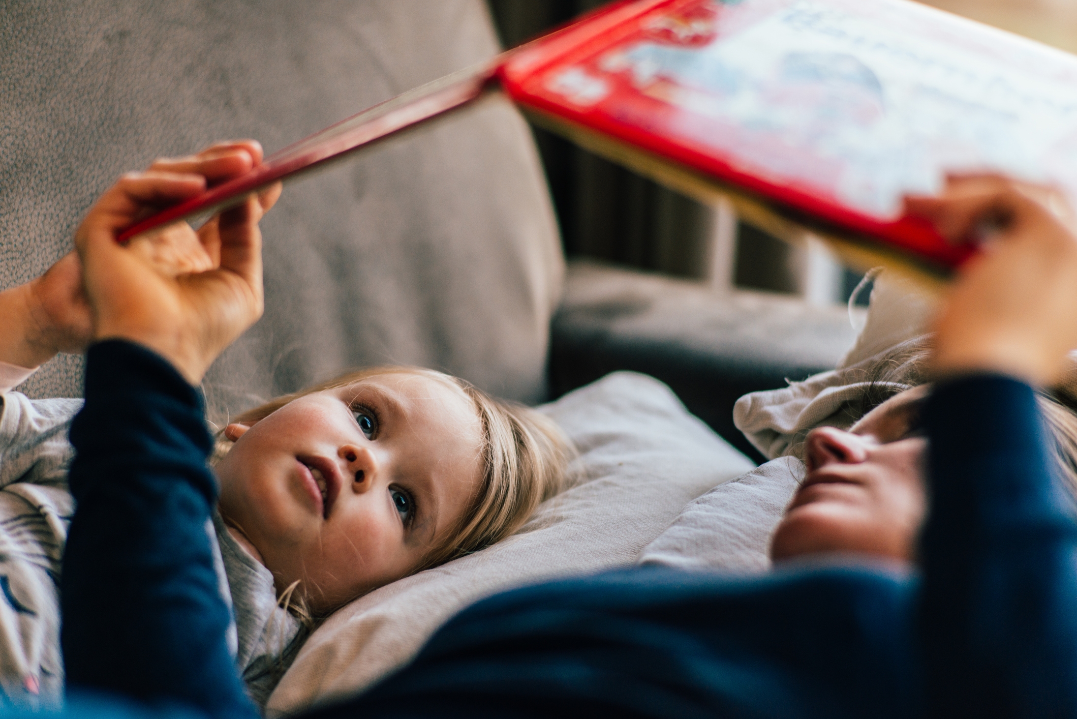 Adult and child lying on a couch, reading a book together, with the child attentively looking at the pages