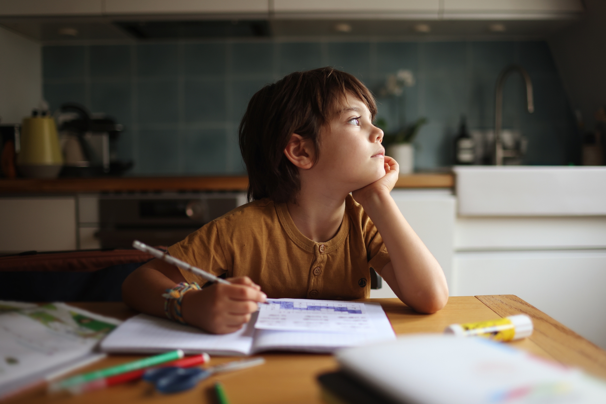Child daydreams while sitting at a table, surrounded by pencils and a drawing book, with a thoughtful expression in a kitchen setting