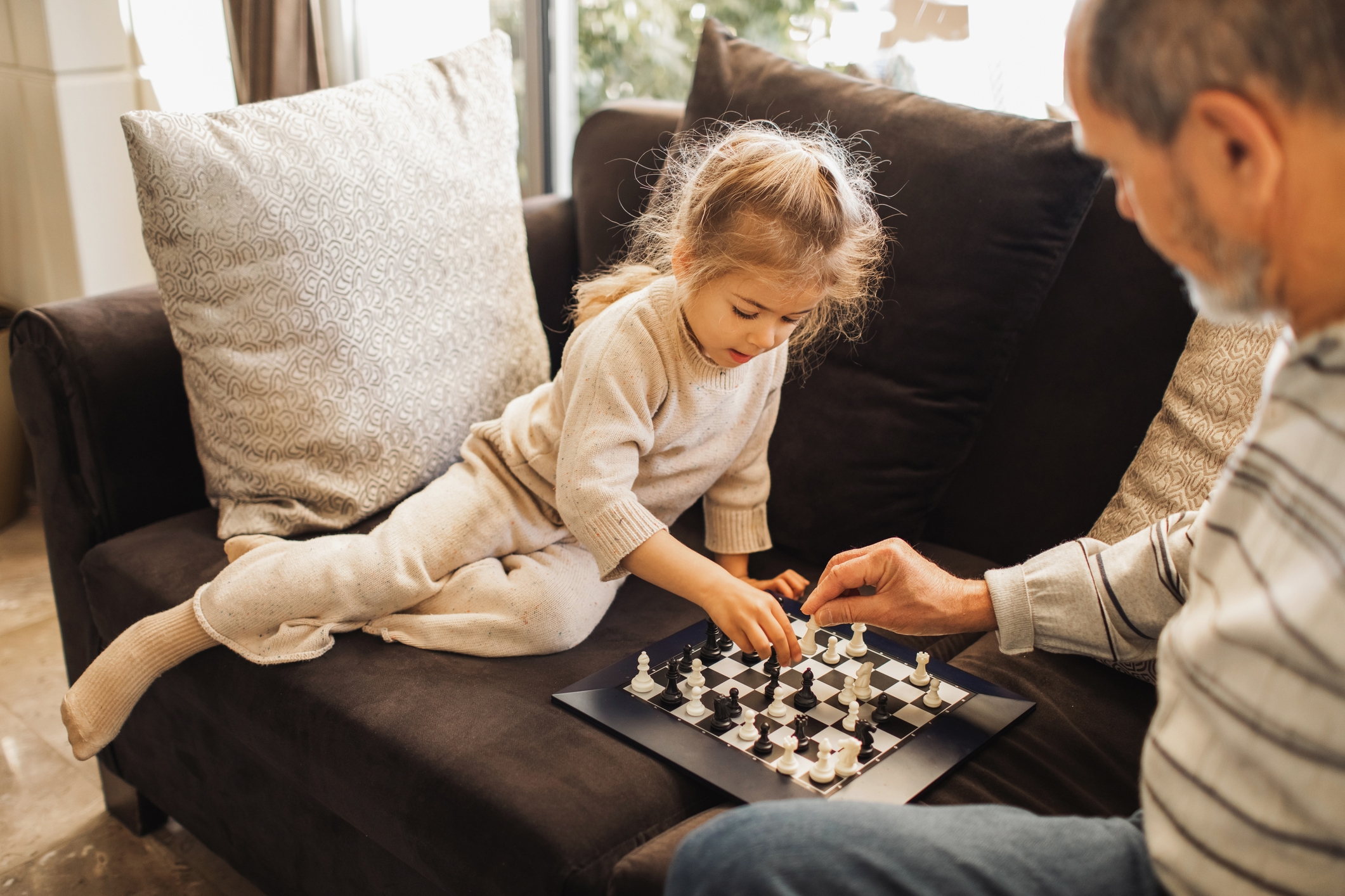 Child and older adult playing chess on a couch, engaged and focused on the game. The child appears to be making a move
