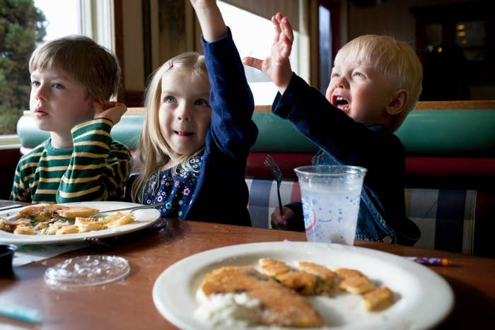 Three children sit at a table with plates of food in front of them, two are eating, and one is raising their hand enthusiastically