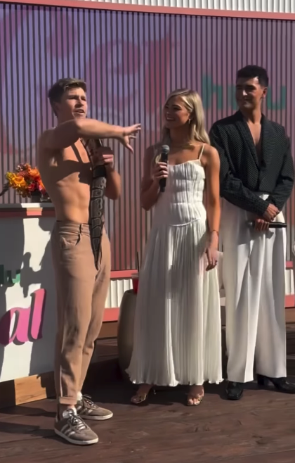 Three people on stage; one shirtless holding a snake, one in a white dress with a microphone, and one in a black shirt and white pants