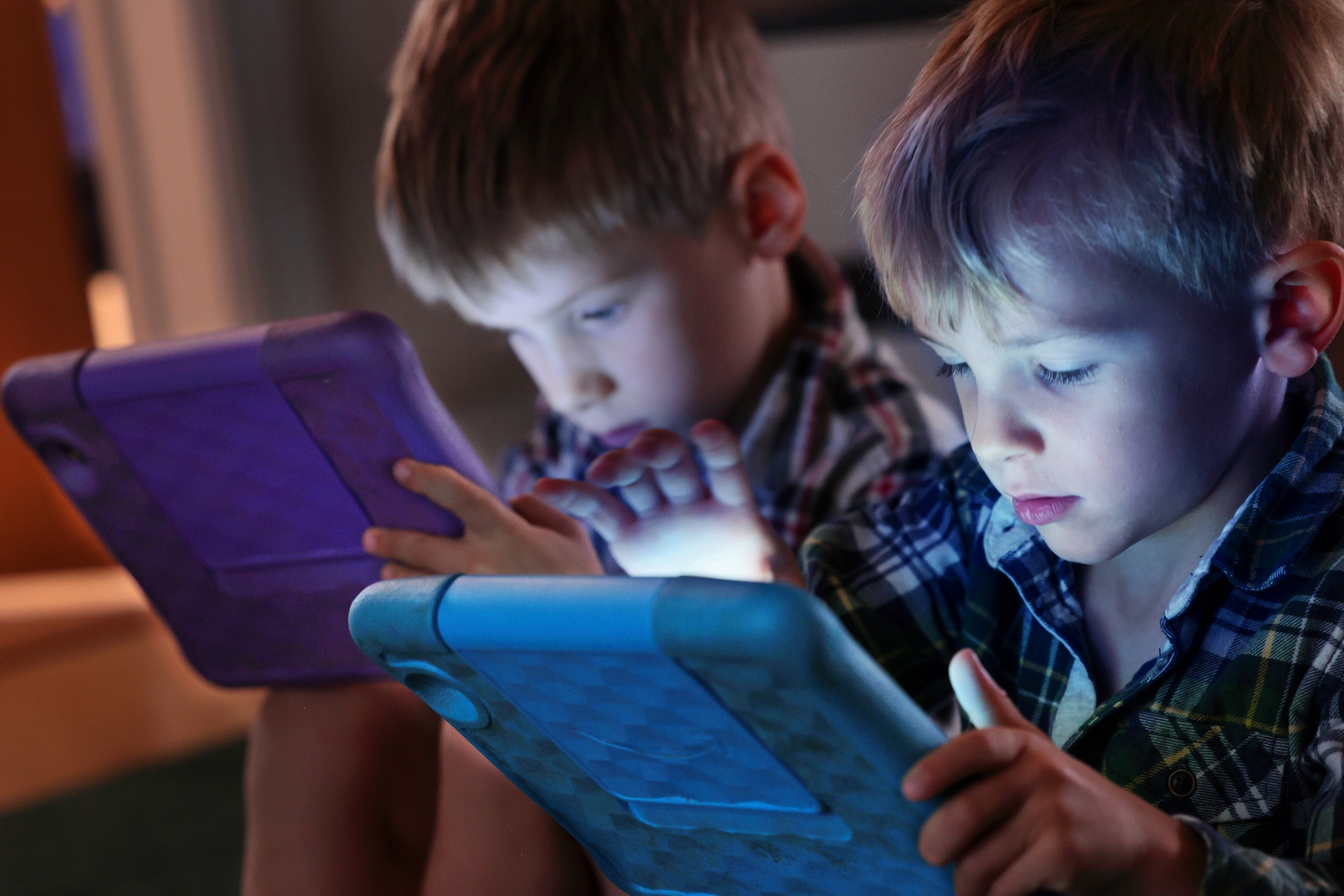 Two young children focus intently on tablets with protective cases, engaging with the screens in a dimly lit room