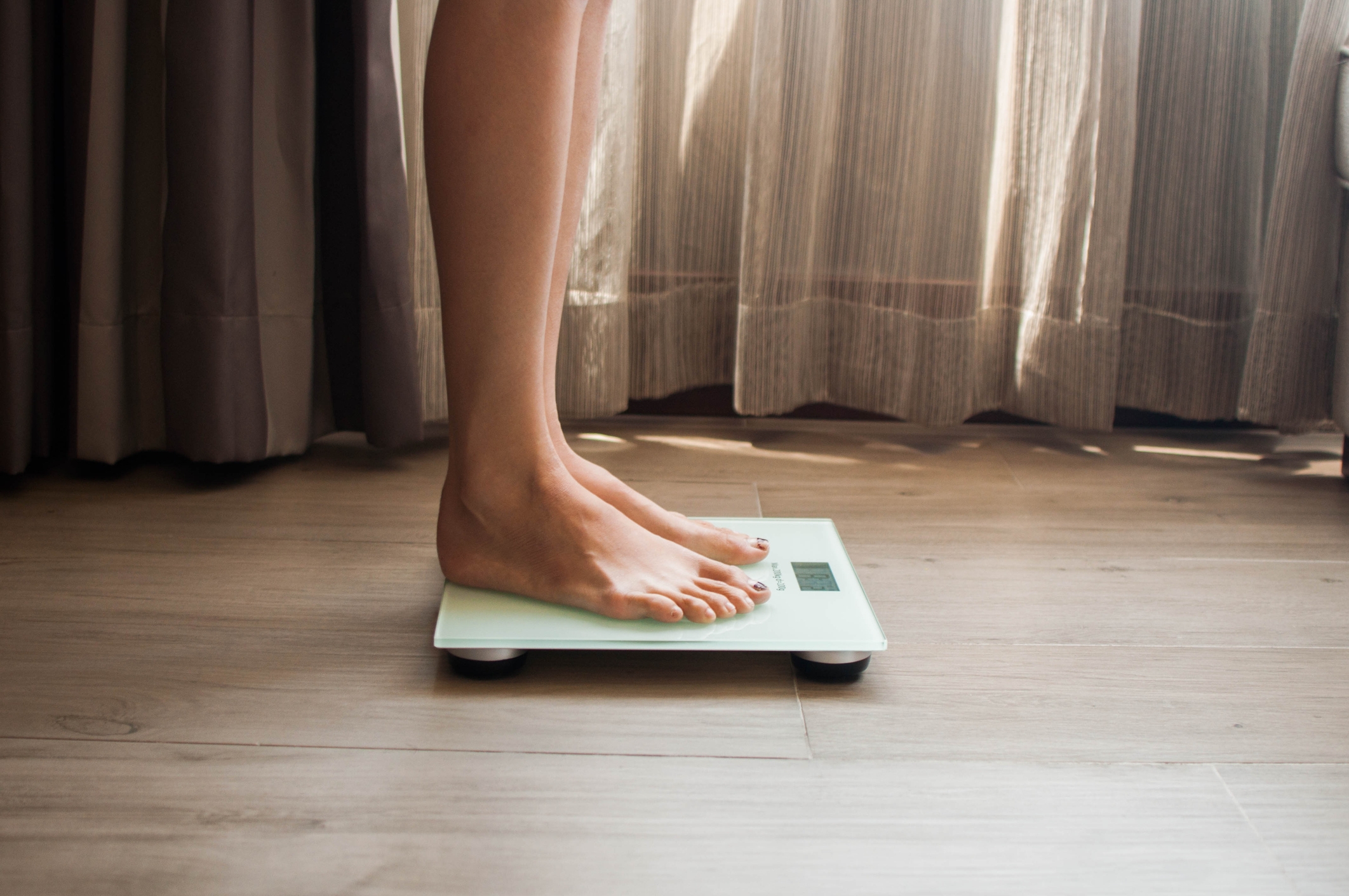 A person standing on a digital scale in a room with curtains, only their legs and feet are visible