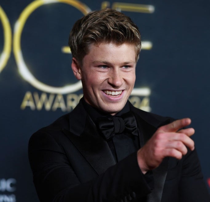 Person in formal black tuxedo smiling and pointing at a media event