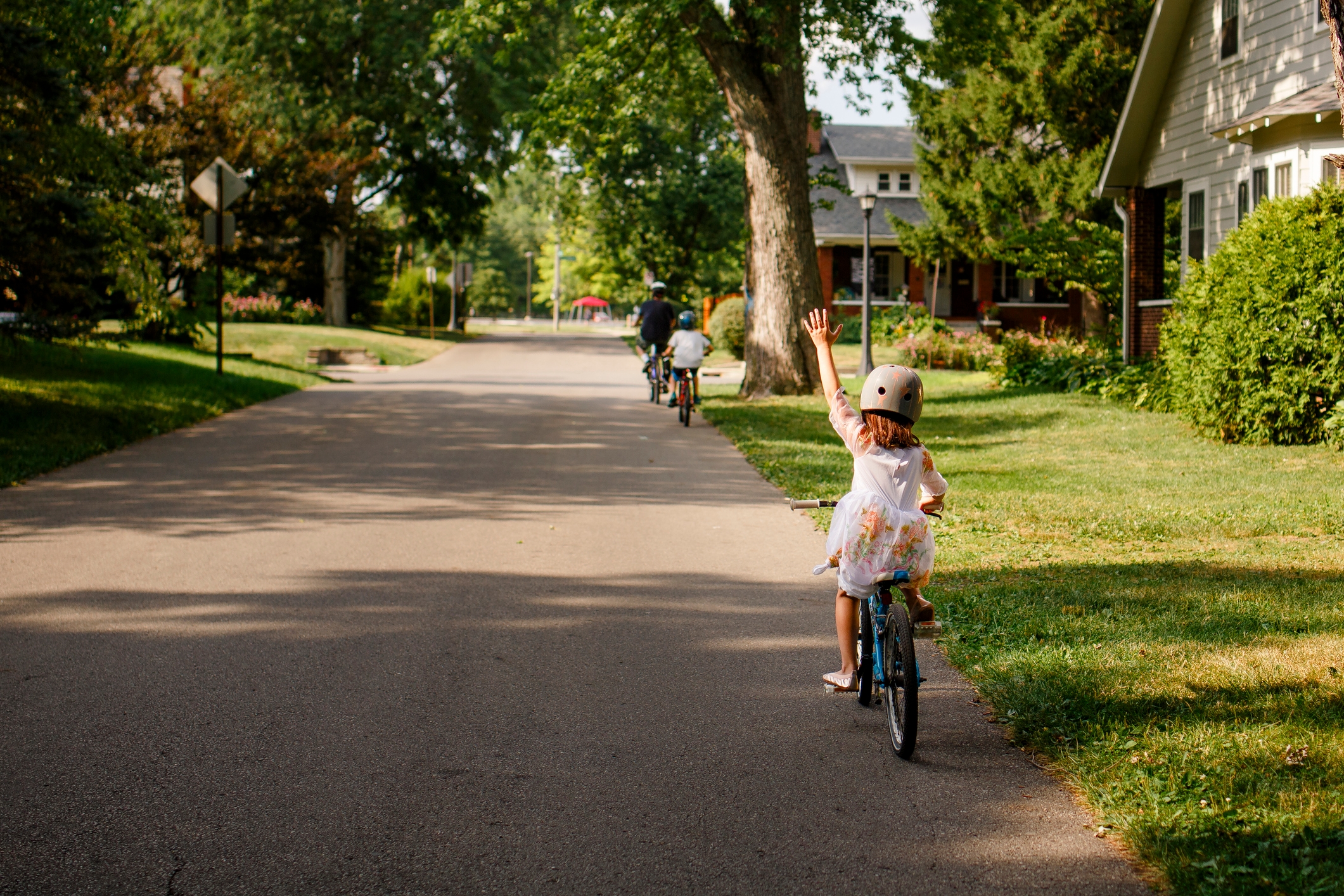 A child on a bicycle waves while riding on a suburban street with two other cyclists ahead. Trees and houses line the quiet road