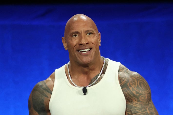 男性がステージに立ち、白いタンクトップとジーンズを着て微笑んでいる。腕にタトゥーがある。