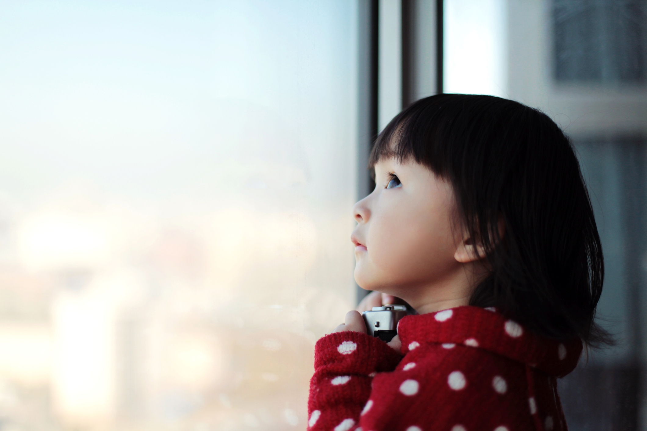 Child in a polka-dot sweater gazes thoughtfully out a window, holding a small object