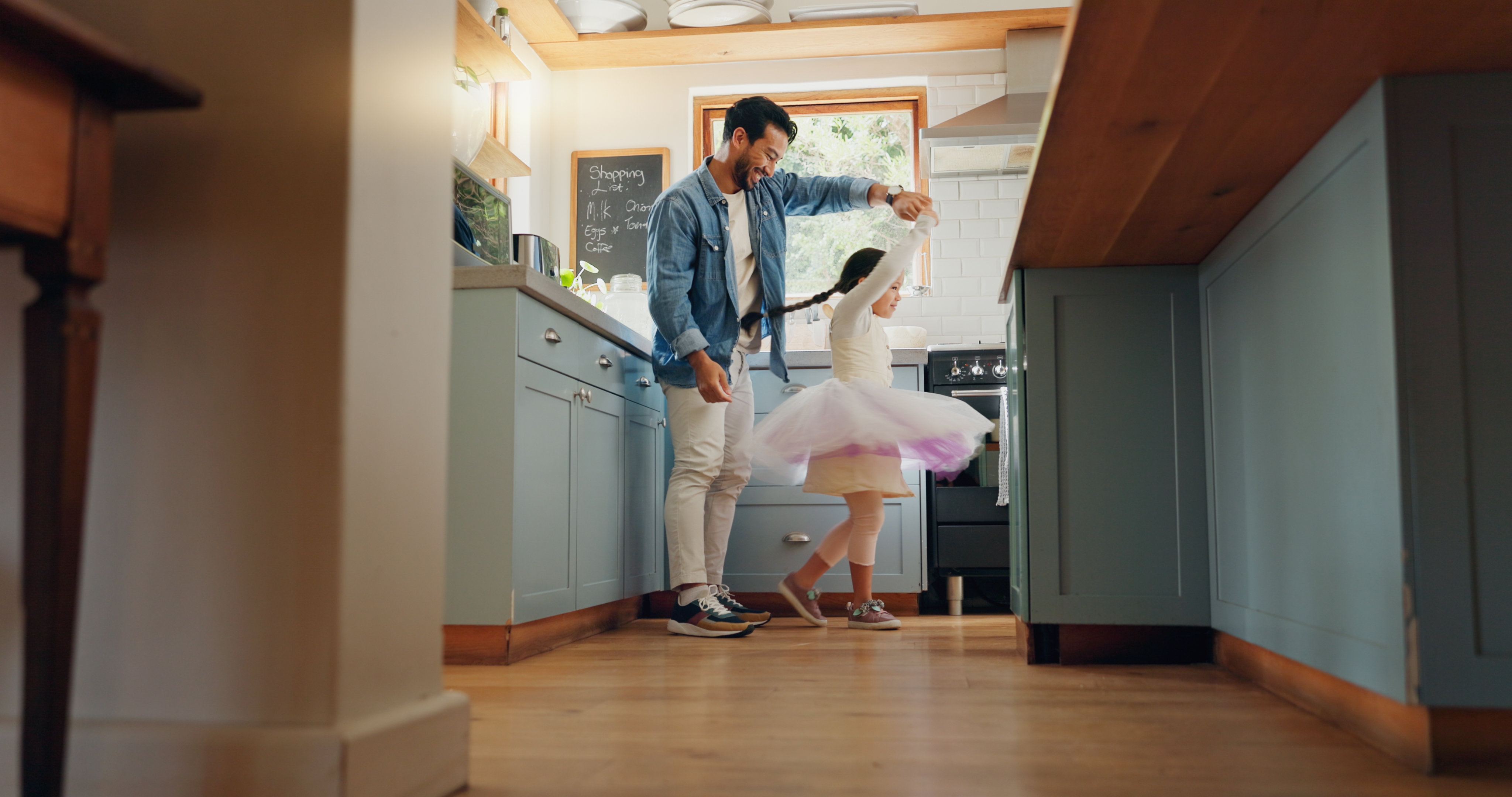 A man dances with a child in a kitchen. The child wears a tutu and spins joyfully, while the man smiles, creating a happy, playful moment