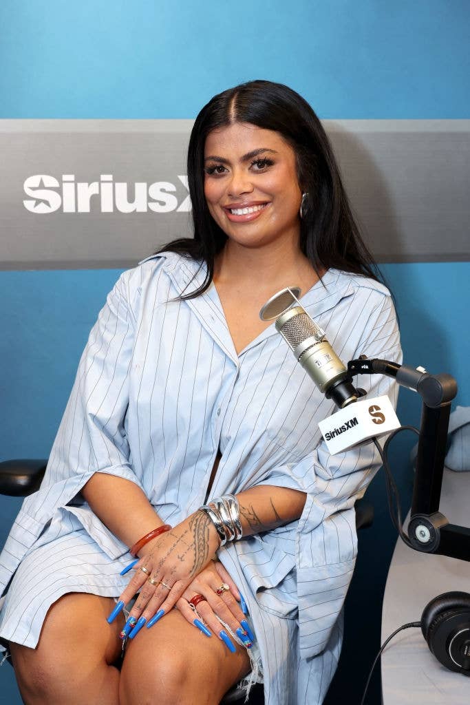 Person in a pinstriped shirt sits in a radio studio with microphone, smiling at the camera and displaying tattooed hands