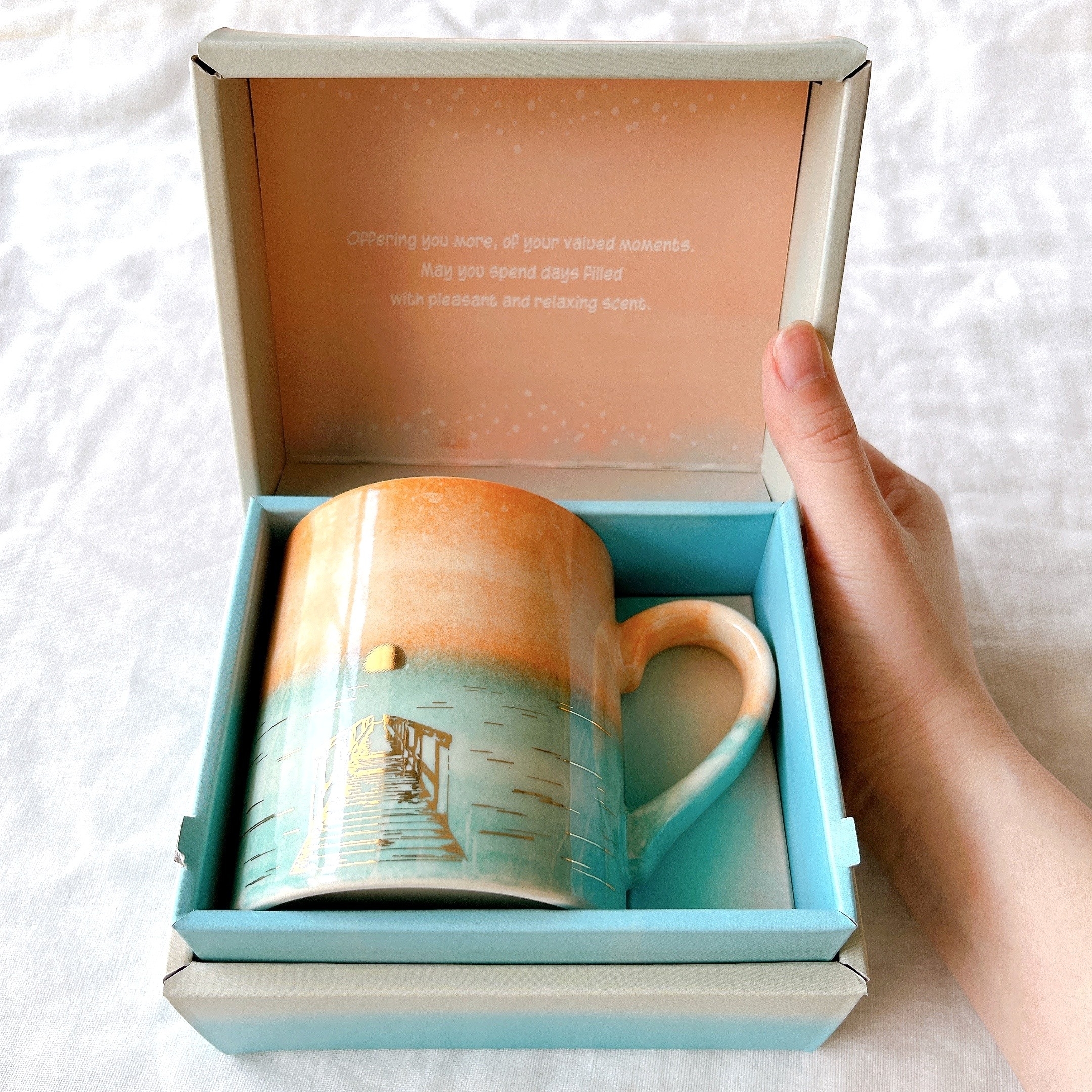 箱に入った風景イラストのマグカップを持つ手。箱内側にメッセージが印刷されています。