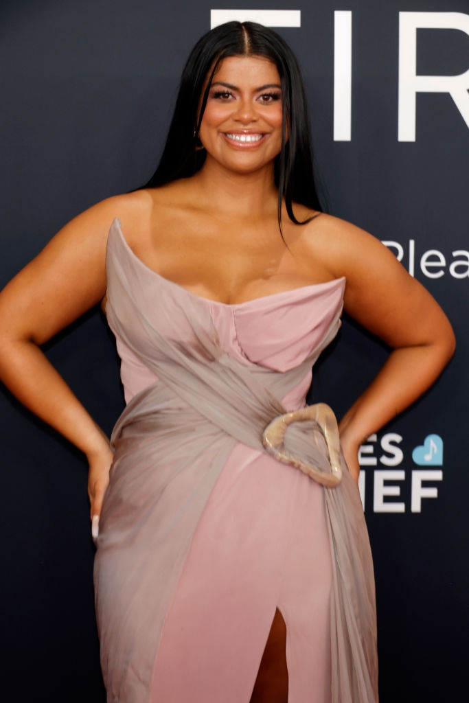 Person on red carpet wearing a stylish off-shoulder gown with a unique belt detail. They are smiling and posing confidently