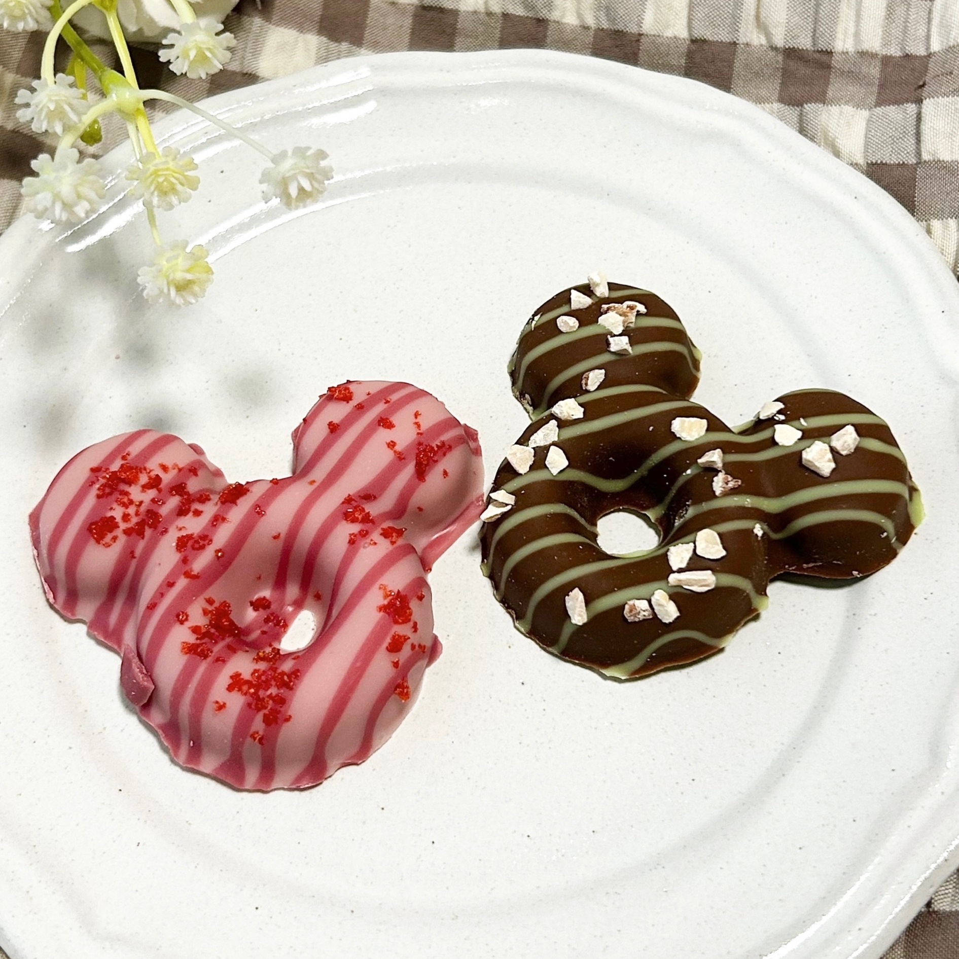 2つのミッキーマウス形のドーナツが白い皿に乗っている。左はピンクのアイシング、右はチョコレートとストライプ模様。