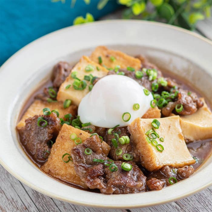 揚げ豆腐と牛肉の煮込み、中央に温泉卵が乗っている料理。上に刻みネギがトッピングされている。