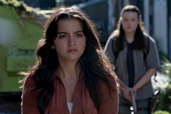 Dos personas en una escena tensa al aire libre en un drama. Una está al frente mirando seria, mientras la otra está detrás sosteniendo un objeto