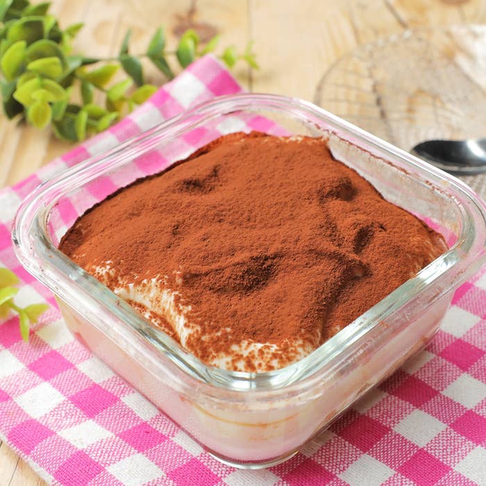 ガラス容器に入ったティラミスが木製テーブルの上に置かれ、周りには緑の植物とピンクチェックの布が見えている。