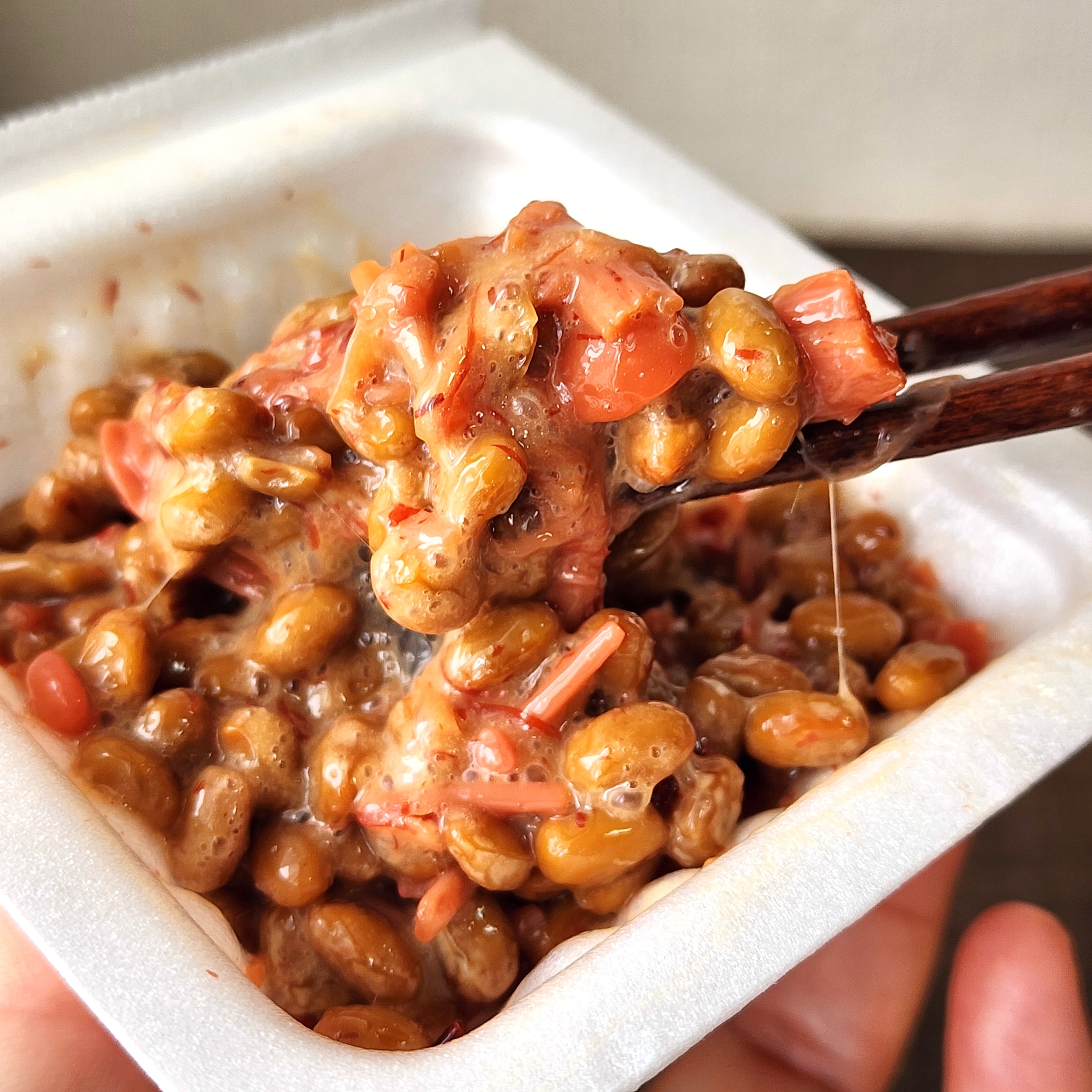 納豆と細切りされたピンク色の具材を箸で持ち上げている様子。泡が立っている。