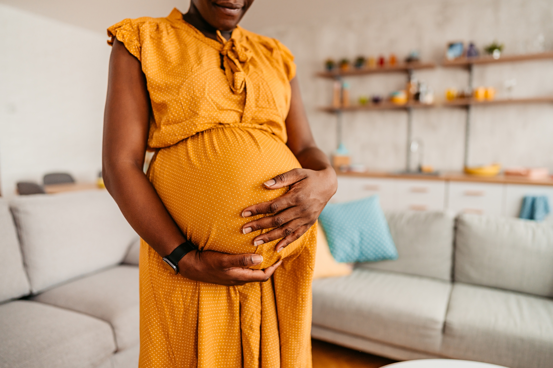 Pregnant person in a cozy living room, gently cradling their belly with hands, wearing a comfortable, sleeveless dress
