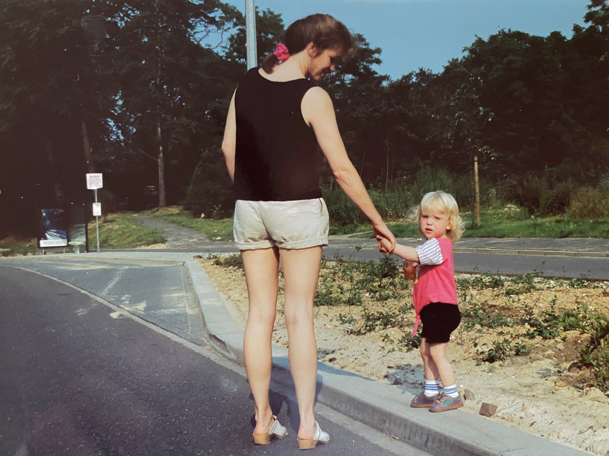 A woman in a sleeveless top and shorts holds hands with a young child on a roadside path, surrounded by trees