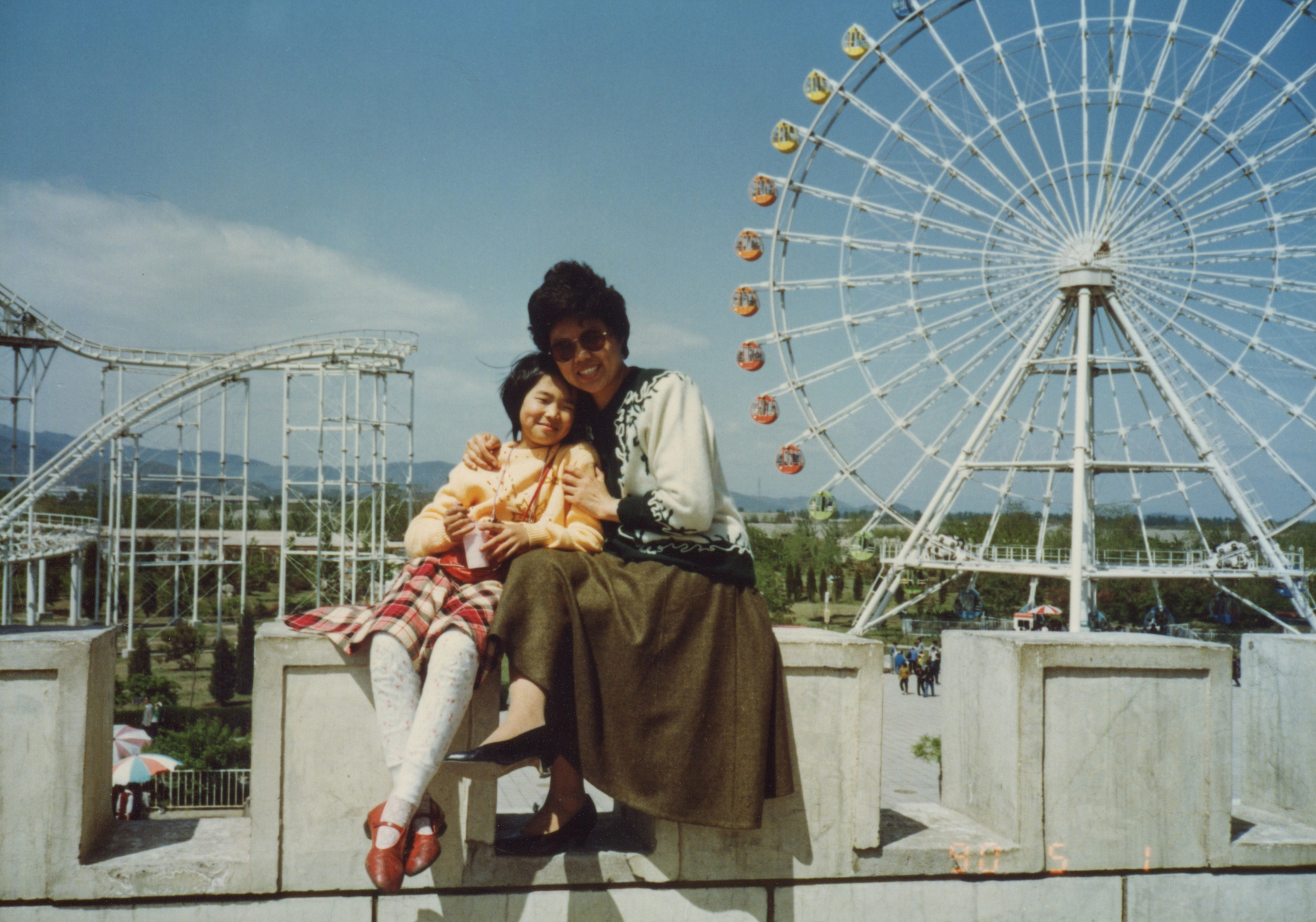 A parent and child sit together smiling in front of a Ferris wheel and roller coaster at an amusement park