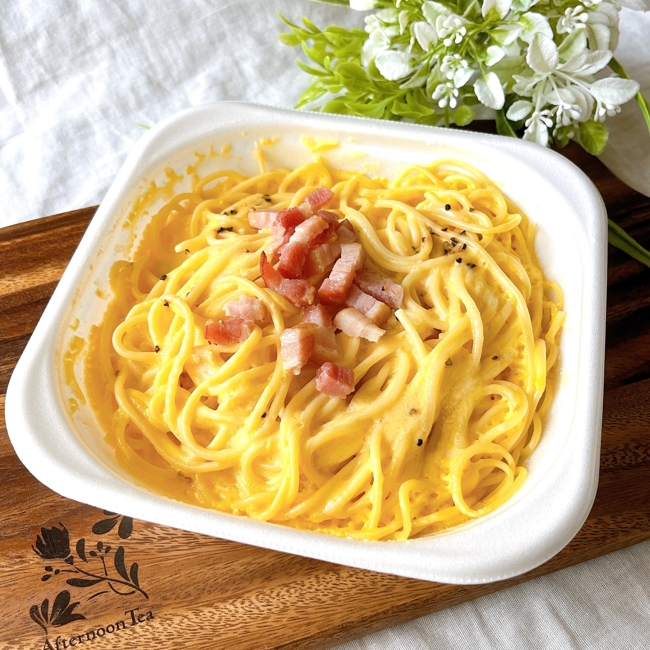 パスタ料理が白い四角い皿に盛られ、ベーコンのトッピングが施されています。背景に白い花。