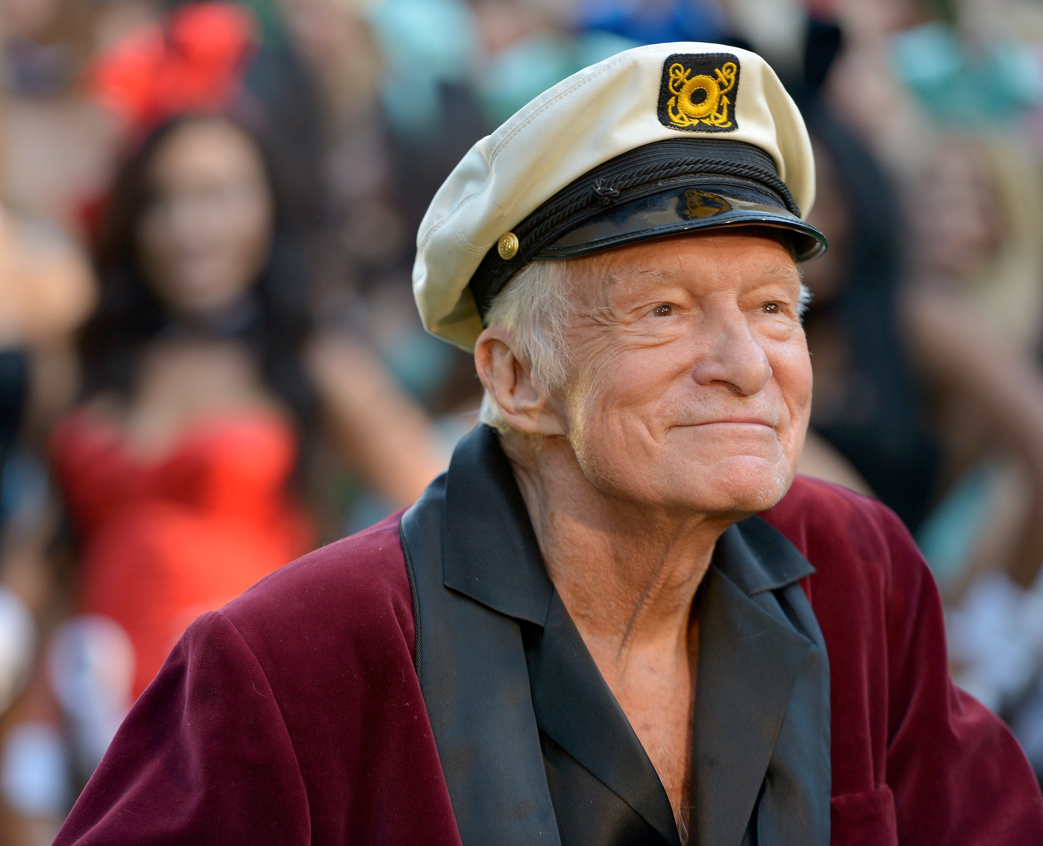 Hugh Hefner, dressed in a velvet smoking jacket and captain's hat, is smiling on a red carpet