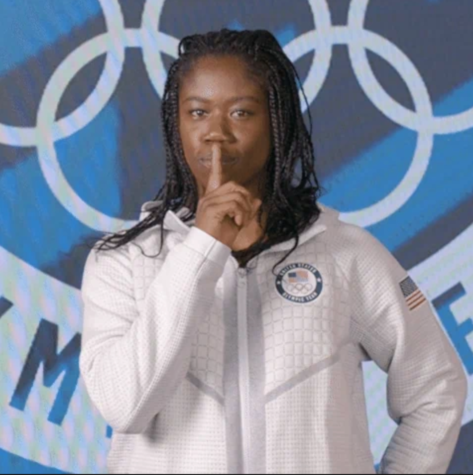 Athlete in a white USA jacket poses with a finger on her lips, standing before Olympic rings backdrop
