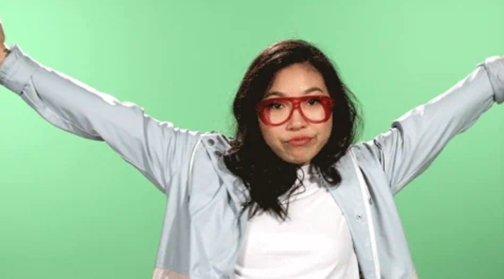 Person with long hair and red glasses poses playfully with arms outstretched against a plain background, wearing a casual jacket and shirt
