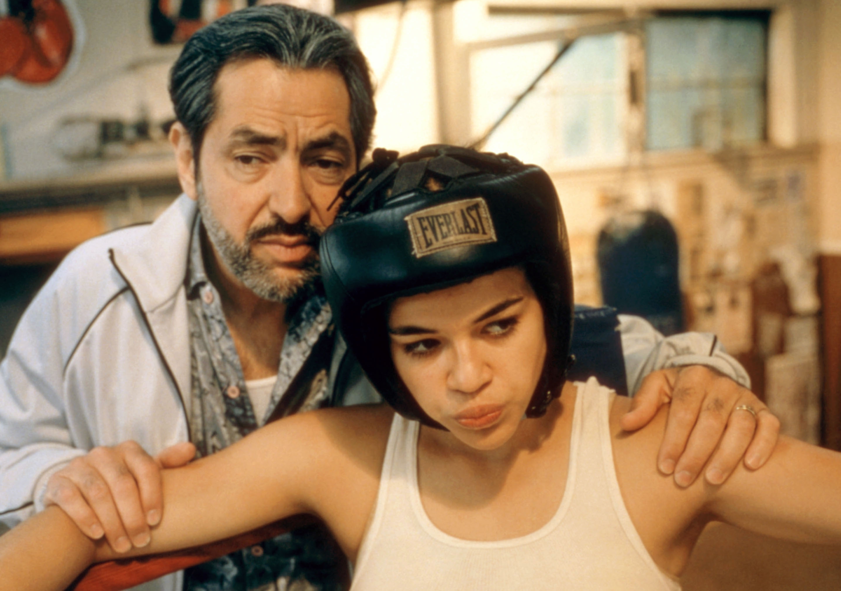 A man coaches a young boxer in a gym. She wears a protective helmet and a tank top, looking focused. He supports her with hands on her shoulders