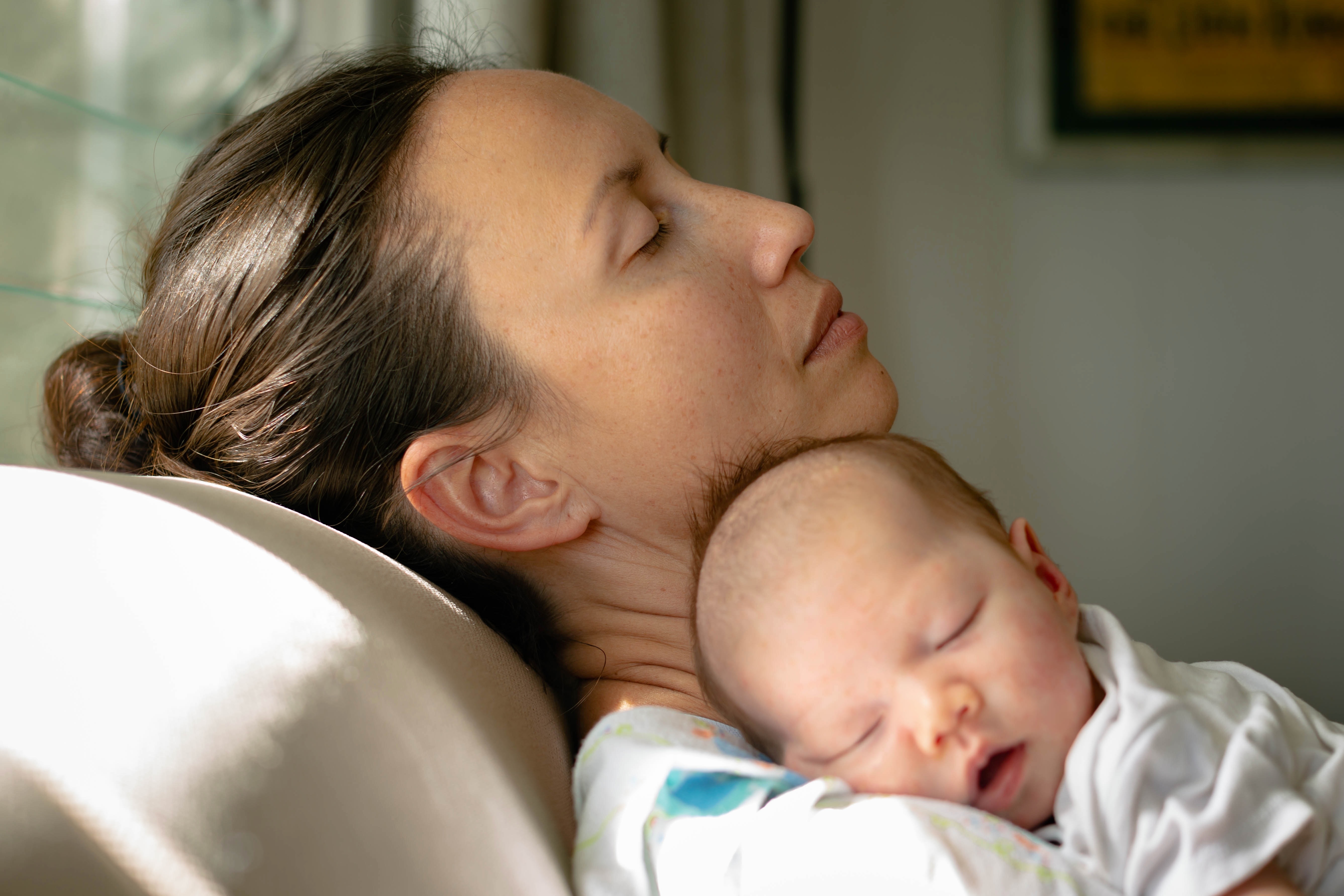 A person rests on a couch with a sleeping baby nestled on their shoulder