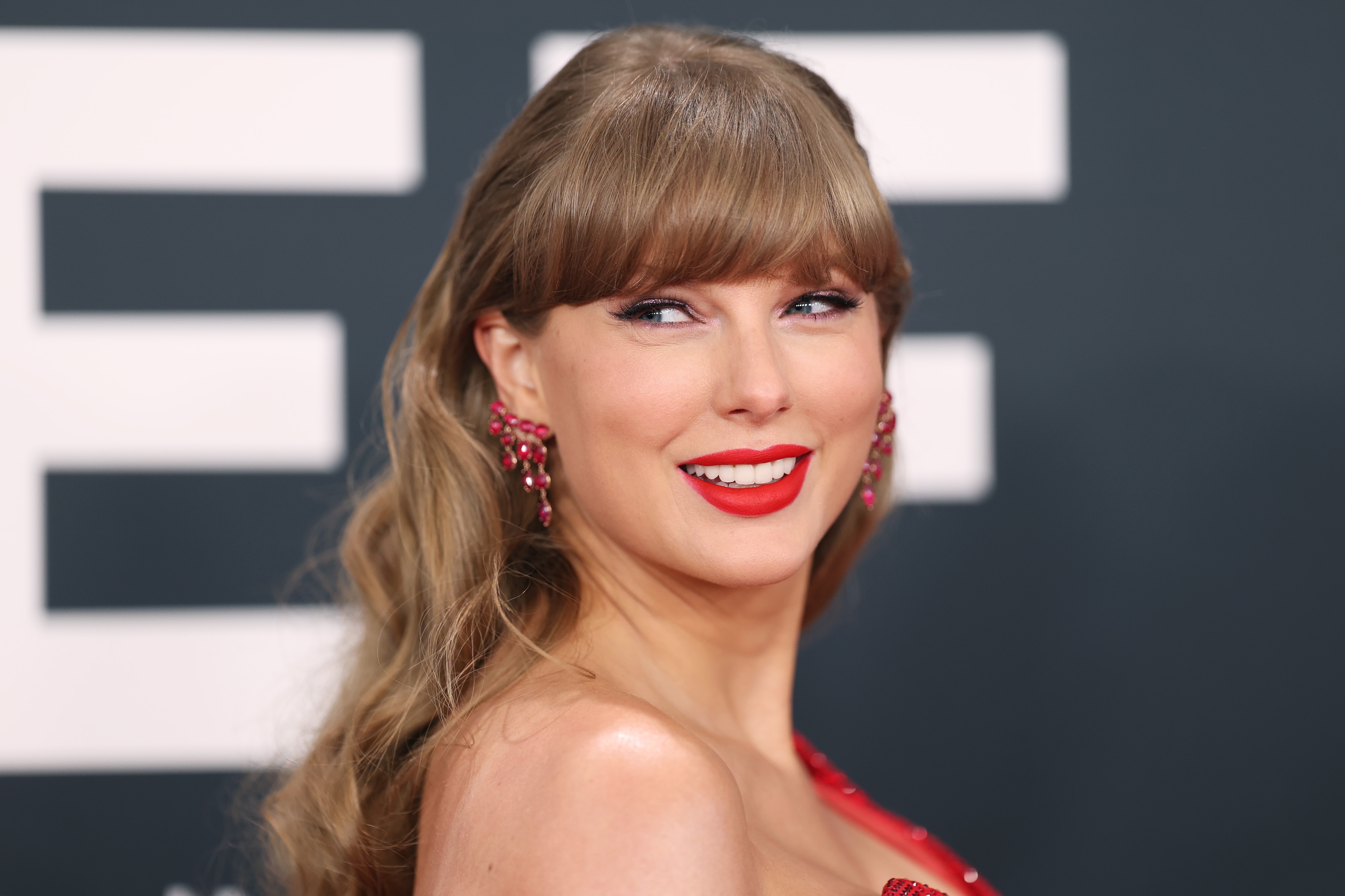 Taylor Swift with long wavy hair and red lipstick smiles while wearing a glamorous dress and statement earrings at an event
