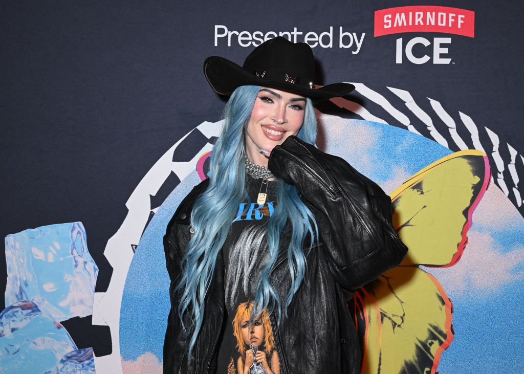 Megan Fox with long blue hair in a black cowboy hat and leather jacket poses at an event backdrop with colorful elements